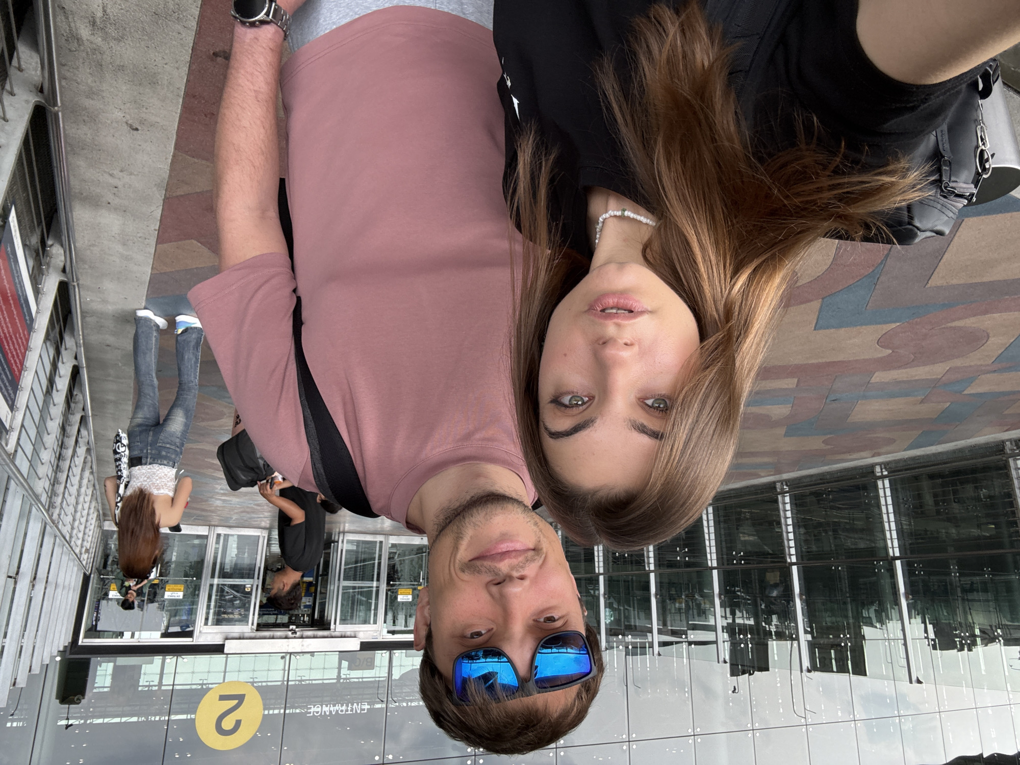 A rotated medium indoor shot depicts a man and a woman posing for a selfie. The man is in the foreground, wearing blue sunglasses and a mauve t-shirt. The woman is next to him, wearing a black shirt and a beaded necklace. Background contains doors and glass facade. The entrance number is indicated as "2". The building's reflection is visible in the glass panes. People are seen walking behind the glass doors. Based on metadata related to the image, the picture was likely taken in Nong Prue Subdistrict, Thailand.