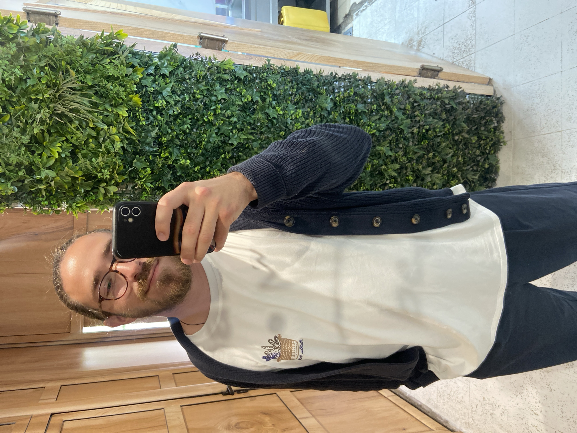 A man wearing glasses and a white t-shirt under a buttoned blue cardigan takes a selfie with a black smartphone in what appears to be an indoor setting. A wall covered in green foliage serves as the backdrop, along with sections of wood paneling. The man is positioned slightly off-center and facing the camera. The flooring appears to be light-colored tiles. There are no explicit signs indicating the location is in Petrovac centar, Montenegro. The setting is neutral.