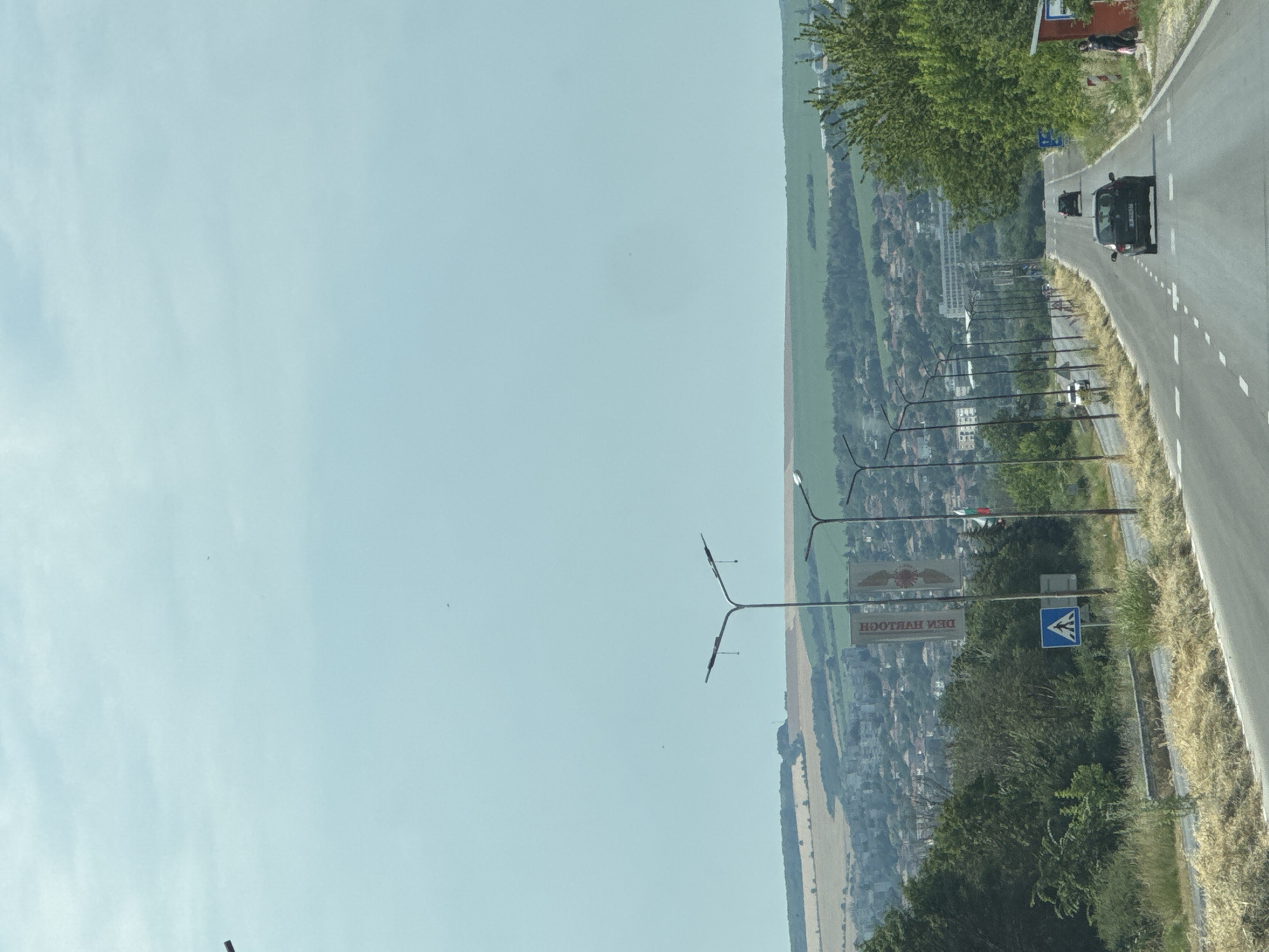 This is a media file captured from a vehicle traveling on a road that curves around a hillside overlooking a town in Kichenitsa, Bulgaria. The scene is bathed in the bright, clear light of what appears to be midday on a sunny day. The sky is a vast expanse of light blue, with a few faint clouds visible.
The road itself is paved and has white dashed lines marking the lanes. There are two dark-colored cars visible on the road, one further ahead than the other. On the left side of the road, there is a steep hillside covered in green fields, trees, and scattered buildings of the town below. The town is densely packed with red-tiled roofs. In the foreground, there are several lampposts and a sign with Cyrillic text that reads "ДЕНИ ХУЛТОЧ" (which can be transliterated as "DENI HULTOCH"). Adjacent to this is a blue sign with a white pedestrian crossing symbol, indicating a crosswalk. Further up the hill, there are more trees and a small red-roofed building. The overall impression is one of a scenic drive through a rural European landscape on a pleasant day.