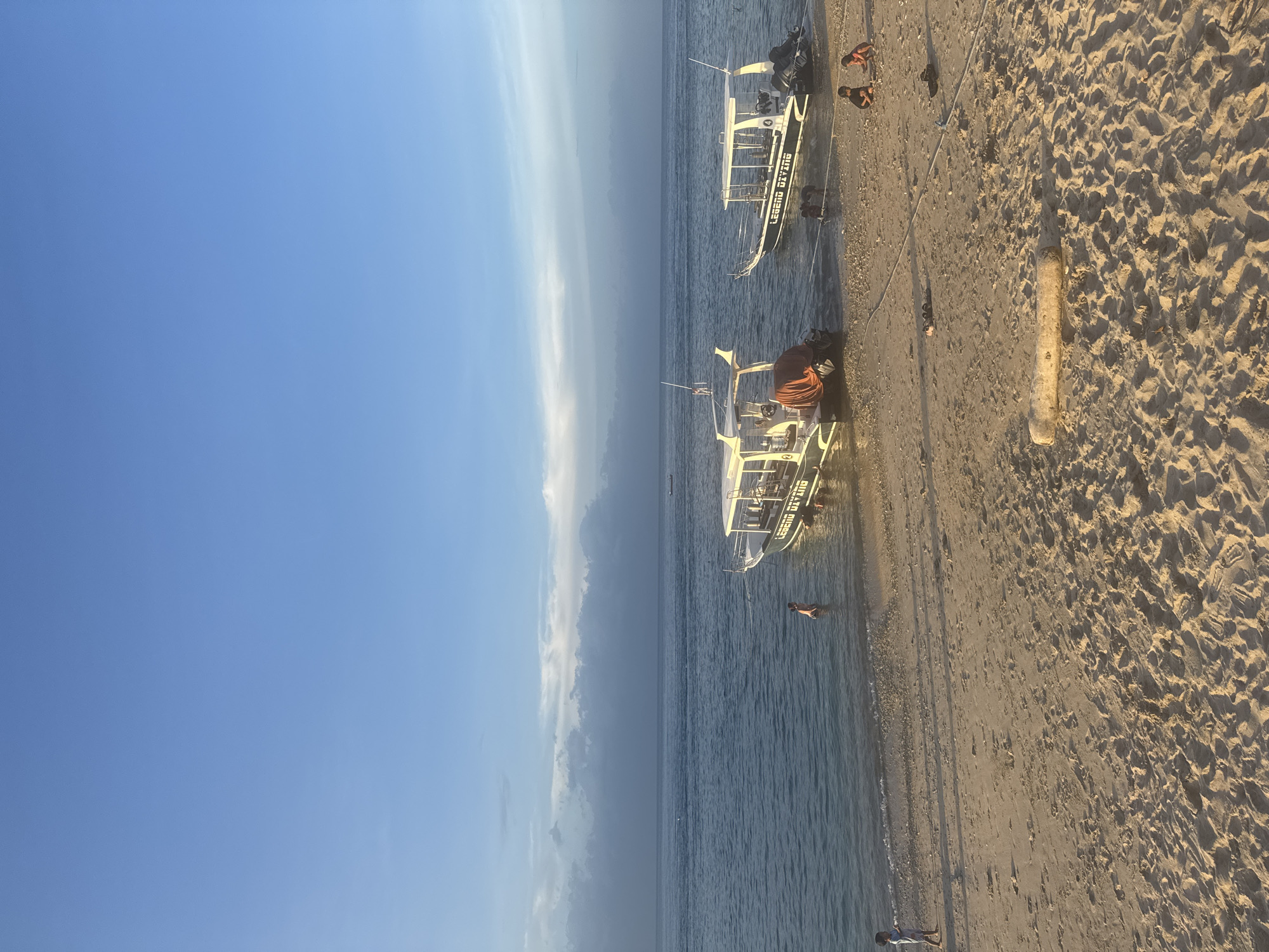 The image depicts a beach scene, possibly in Nusa Penida, Indonesia, given the boat name "LEGEND DIVING" visible on the boats, which is likely a diving company operating in the area. Two boats are docked or beached close to each other on the sandy shore, with people around and on them. A larger cluster of people are on the beach, with some moving around. The beach is sandy, and the water is calm. The sky is clear blue, with some clouds on the horizon.