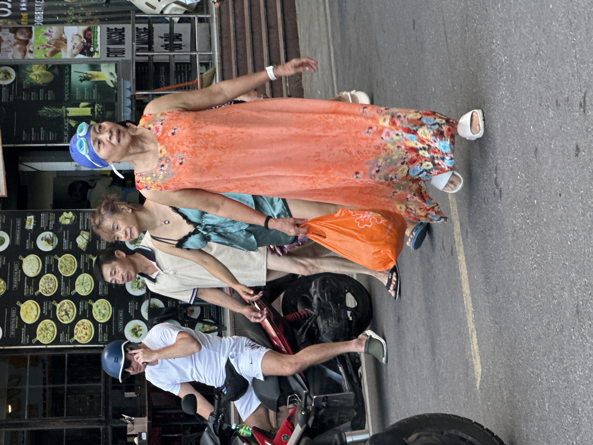 The image shows a scene in Bac Nha Trang Ward, Vietnam. Four people are walking on the sidewalk next to a street with a parked scooter. An older woman is wearing a peach-colored dress, a blue swim cap, and goggles. Behind her are a younger woman in a blue dress and a man wearing a tan shirt. In the foreground, a man wearing a white shirt and shorts is standing next to a red and black scooter, holding a blue helmet. A restaurant or shop is visible in the background, with signs promoting food, massage, and drinks.