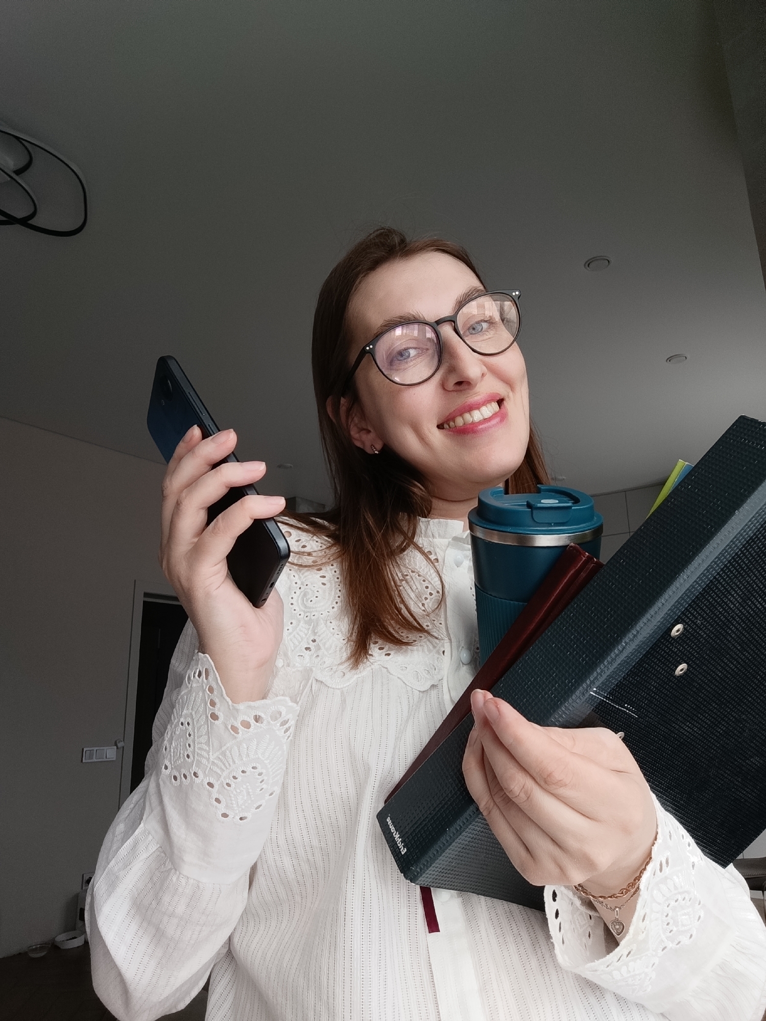 A young woman, likely in her 20s or 30s, smiles broadly at the camera in a selfie-style shot taken indoors in Chaikovsky, Russia. She has light brown hair and wears black-framed glasses, revealing prominent dimples. Her expression is cheerful and positive.
She is dressed in a white long-sleeved blouse, featuring delicate lace and embroidered patterns on the collar and cuffs. Her hands are full, indicating a state of busyness or readiness. In her left hand, she holds a black smartphone. Her right arm and hand support a collection of items: a teal and silver reusable coffee cup, a smaller dark red book or folder, and a large dark blue or black lever arch binder. A gold chain bracelet with a heart-shaped charm adorns her right wrist.
The background is a simple, light grey wall or ceiling, with a few indistinct elements that appear to be indoor fixtures. The overall scene suggests she is prepared for a day of work or study. The only legible text is the partially obscured brand name "Евроканц" (Evrokanz) on the spine of the large binder. The indoor lighting does not indicate a specific time of day.