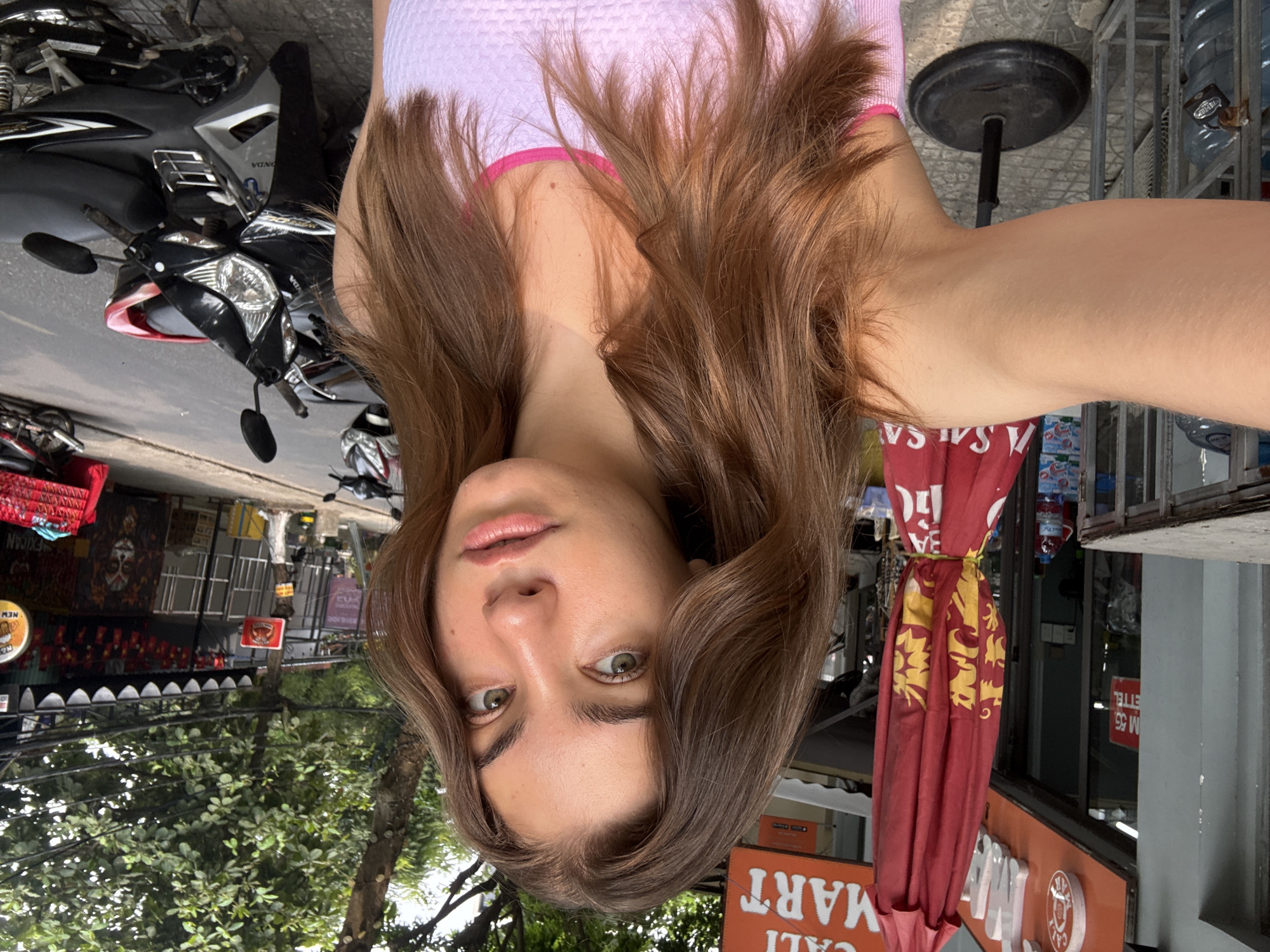 The image is rotated 90 degrees clockwise, depicting a young woman taking a selfie in what appears to be an urban street or sidewalk in Da Nang, Vietnam. She is the central subject, looking directly at the camera with a neutral expression. She has shoulder-length light brown hair, which frames her face and spreads around her shoulders, and distinct green eyes. She is wearing a light pink or purple sleeveless top with a darker pink trim.
The background reveals a bright, daytime street scene. To the left of the woman, several motorcycles are parked, including a black Honda with "HONDA" and "VONG" visible on its body. Further back, green trees and multi-story buildings with various shops and awnings are visible. One shop front in the distance has text that includes "NEW MEXICAN". A red woven basket is also seen on the far left.
To the right, a red fabric umbrella or awning, partially rolled up, displays yellow patterns and the word "SALSA" in white. Below this, an orange sign prominently features "CALI MART" in white lettering. Behind the umbrella and signs, there's a glimpse of a shop interior with racks holding large water bottles. A black, circular base of a stand is also present near her right shoulder. Other text, such as "ETEL" and "M 55" (reversed), can be seen on signs or reflections in the background. The setting suggests a lively commercial area in a Southeast Asian city.
