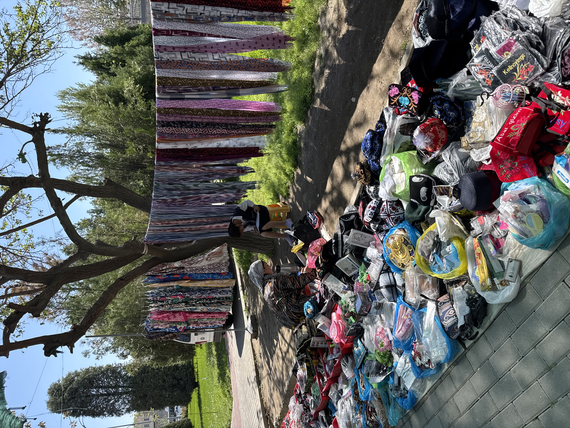 The image shows an outdoor market scene in Samarkand City, Uzbekistan. A vendor wearing a headscarf sits on the ground beside a large assortment of merchandise, primarily hats and textiles. These items are displayed on a blanket or mat on the paving stones and include many colourful hats. Rolls of fabric hang vertically, suspended from a frame attached to a tree branch. Another person sits on a small stool near the fabric. The background includes trees, grass, and a partial view of a light-coloured building. The day appears sunny.