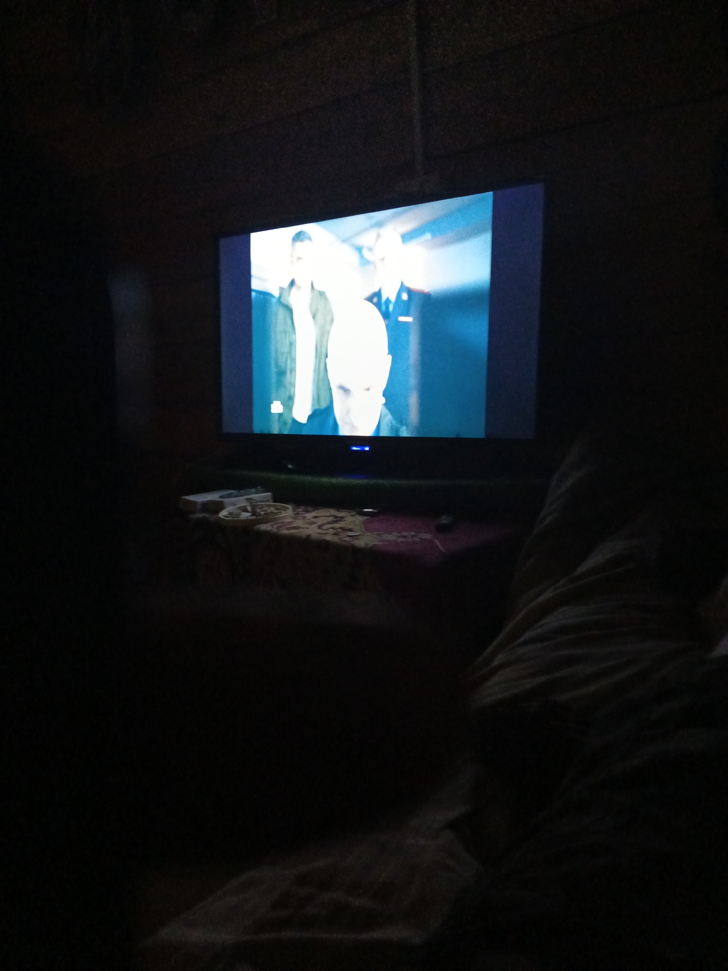The media file depicts a dimly lit room where a television is displaying a scene with three individuals. Two people are partially visible on the left of the screen, while a third person, a man with a bald head, is prominently featured in the center, looking directly at the camera. The television screen is the primary source of light in the room, casting a blueish glow.
On a table in front of the television, several objects are visible, including what appears to be a white rectangular object, possibly a remote or a game controller, and a circular object with a patterned surface. The table is covered with a patterned cloth.
The foreground of the image is very dark, obscuring most of the room and any potential viewers. The impression is of someone or people watching television in a dark environment, possibly at night. The overall mood is quiet and relaxed, suggesting a casual viewing experience. No specific weather conditions are discernible, and the location context of Strelka, Russia, does not provide further visual clues within the image itself. No visible text is present in the media file.