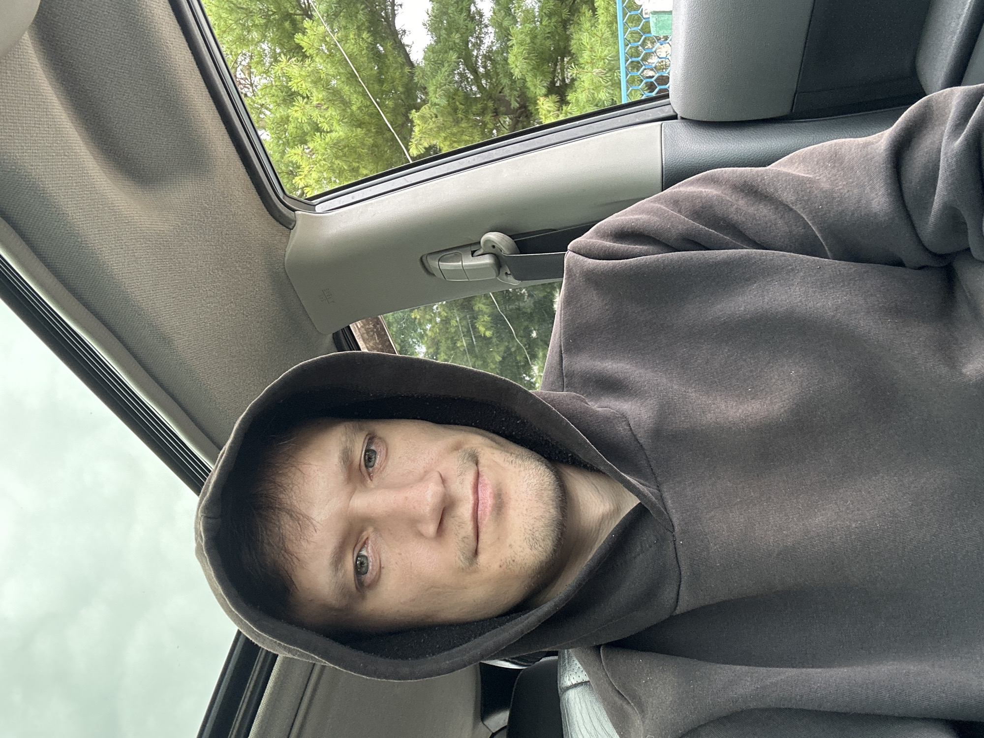 A man wearing a dark hooded sweatshirt is seated inside a vehicle, with the sunroof open. He is looking directly at the camera, his expression neutral. The interior of the car is visible, including a seatbelt mechanism on the right side. Through the window, lush green trees and a blue-railed fence are seen, suggesting an outdoor environment. The sky is overcast. There is no indication of the specific location within Makiv, Ukraine.