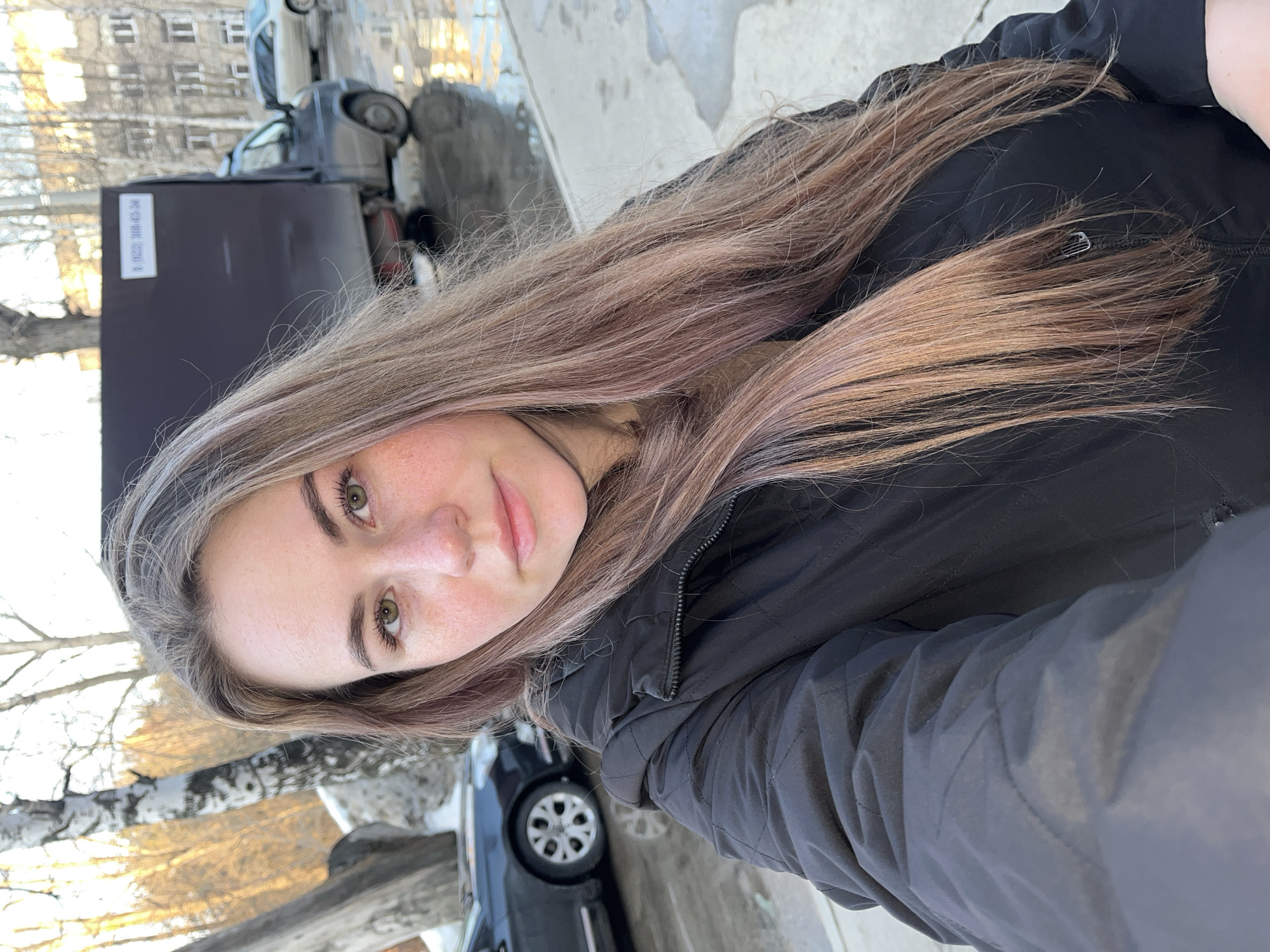 A young adult female is centered in the foreground, taking a selfie outdoors in the city of Chaikovsky, Russia. She has long, light brown hair with lighter, possibly pinkish, streaks, green eyes, and freckles visible on her nose and cheeks. She is wearing a black, quilted jacket. Her expression is neutral, and she is looking directly at the camera.
The immediate ground is wet, dark pavement reflecting light. To the left, two birch trees with white bark stand near a patch of snow. A dark-colored sedan is partially visible behind the trees. To the right, a dark box truck, possibly a GAZelle model, is parked on the wet pavement. A white rectangular sticker with the black text "(8422) 306-62-04" is affixed to the rear of the truck's cargo box. In the mid-ground, beyond the truck, a multi-story residential building with multiple windows is visible. Further to the left in the background, a wooded area with bare trees is illuminated by warm sunlight, suggesting either early morning or late afternoon. The overall lighting is natural and bright.