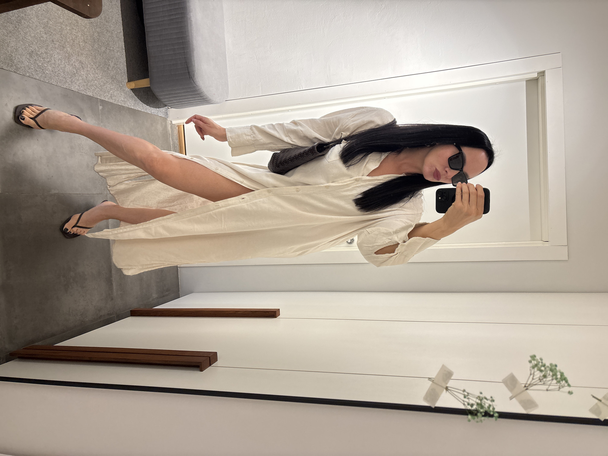 A woman with long black hair wearing sunglasses and a white shirt dress is taking a selfie in front of a mirror. She is holding a black smartphone and has a black bag over her shoulder. She is wearing black flip-flops. The background includes a white wall, a gray couch, and a reflection of decorative floral elements on the mirror. The location is most likely an interior space, possibly a room or apartment, but the city is not possible to infer from the provided image.