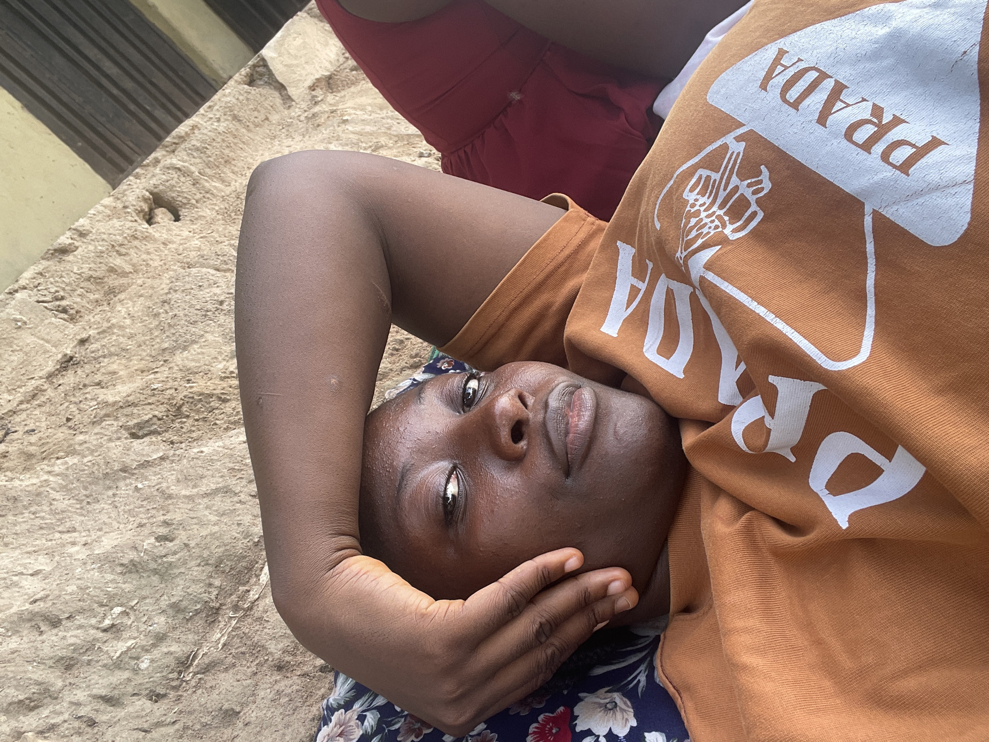 A woman with dark skin and short hair is lying on a concrete surface in Akwanga, Nigeria. She is wearing a brown shirt with "PRADA" written in white letters on it. Her left arm is raised and bent at the elbow, with her hand touching her cheek. She is lying on a patterned cloth, possibly a skirt or blanket. Another person wearing a red dress is visible in the upper center of the image. The background includes a wall or structure with horizontal lines. The lighting suggests it is daytime.