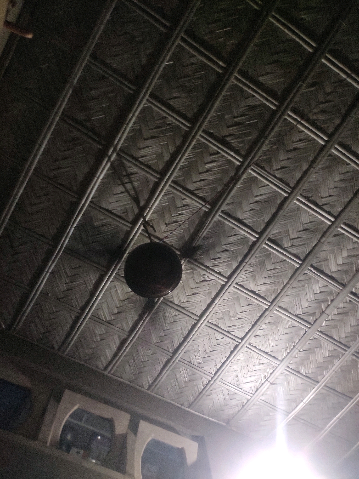 The image shows an interior ceiling constructed from interwoven bamboo, with diagonal bamboo poles forming a criss-cross pattern. A circular, dark-colored object, possibly a light fixture or fan, hangs from the ceiling in the center of the frame. The lighting is dim, with a bright light source emanating from the lower right corner, creating a lens flare effect and casting shadows on the bamboo ceiling. In the lower portion of the image, a portion of a wall with arched openings is visible. The scene appears to be indoors, in a space with a traditional or rustic architectural style.