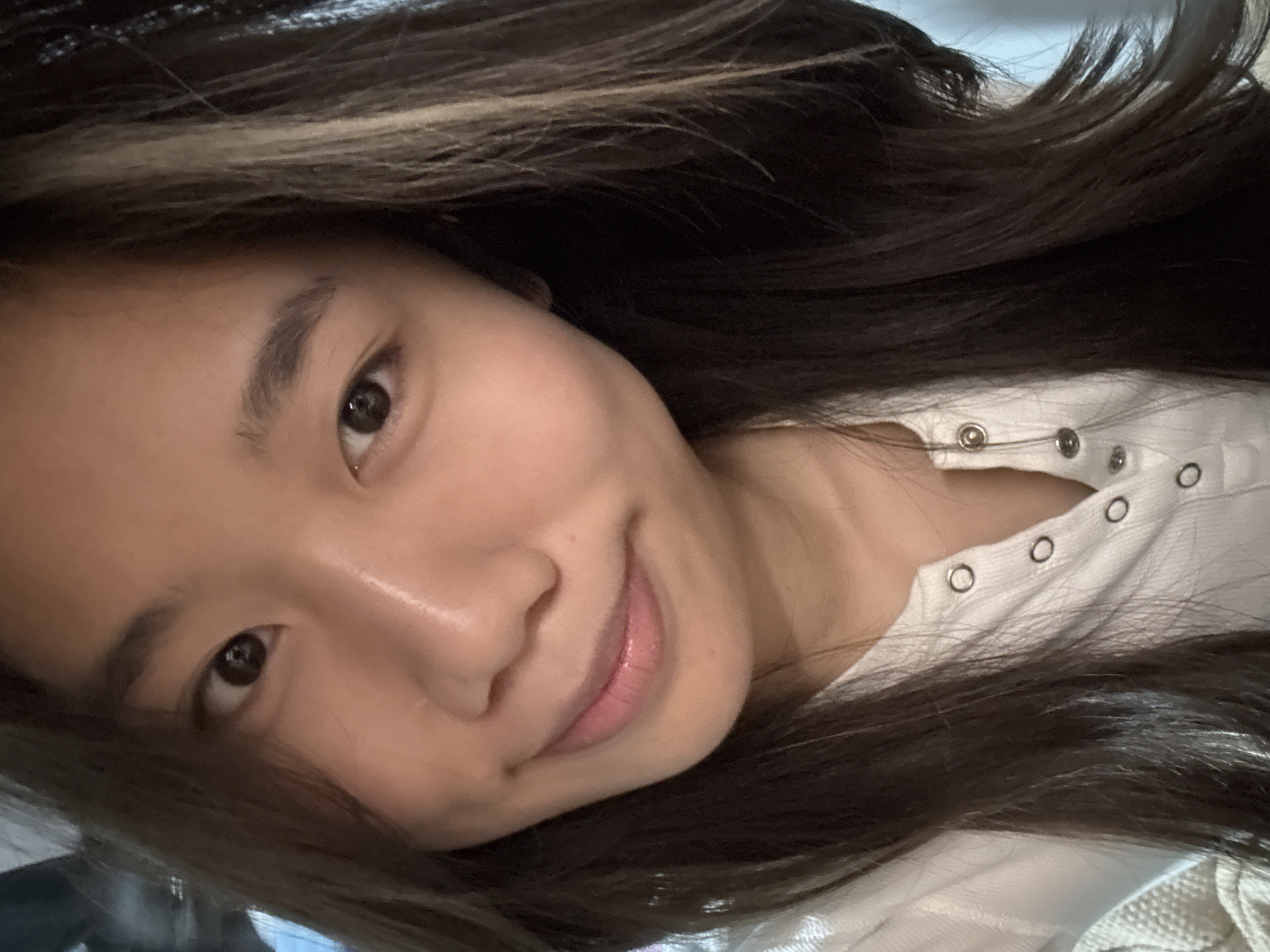 A portrait depicts a single female individual, located in Nan'an District, China. The subject is East Asian, with long, dark brown hair that includes lighter brown or blonde highlights, particularly on the viewer's left side. Her dark brown eyes are open and directly focused forward. A subtle, closed-mouth smile is present, with lips appearing to have a light pink cosmetic application. Her skin tone is light-to-medium, exhibiting some natural sheen on the forehead and nose bridge. She is dressed in a white, ribbed garment featuring a collar and multiple silver snap buttons down the front, with at least two buttons fastened at the neckline. The background is uniformly dark and indistinct, indicating an indoor setting without identifiable features. Illumination is soft, appearing to originate from the subject's upper-left.
