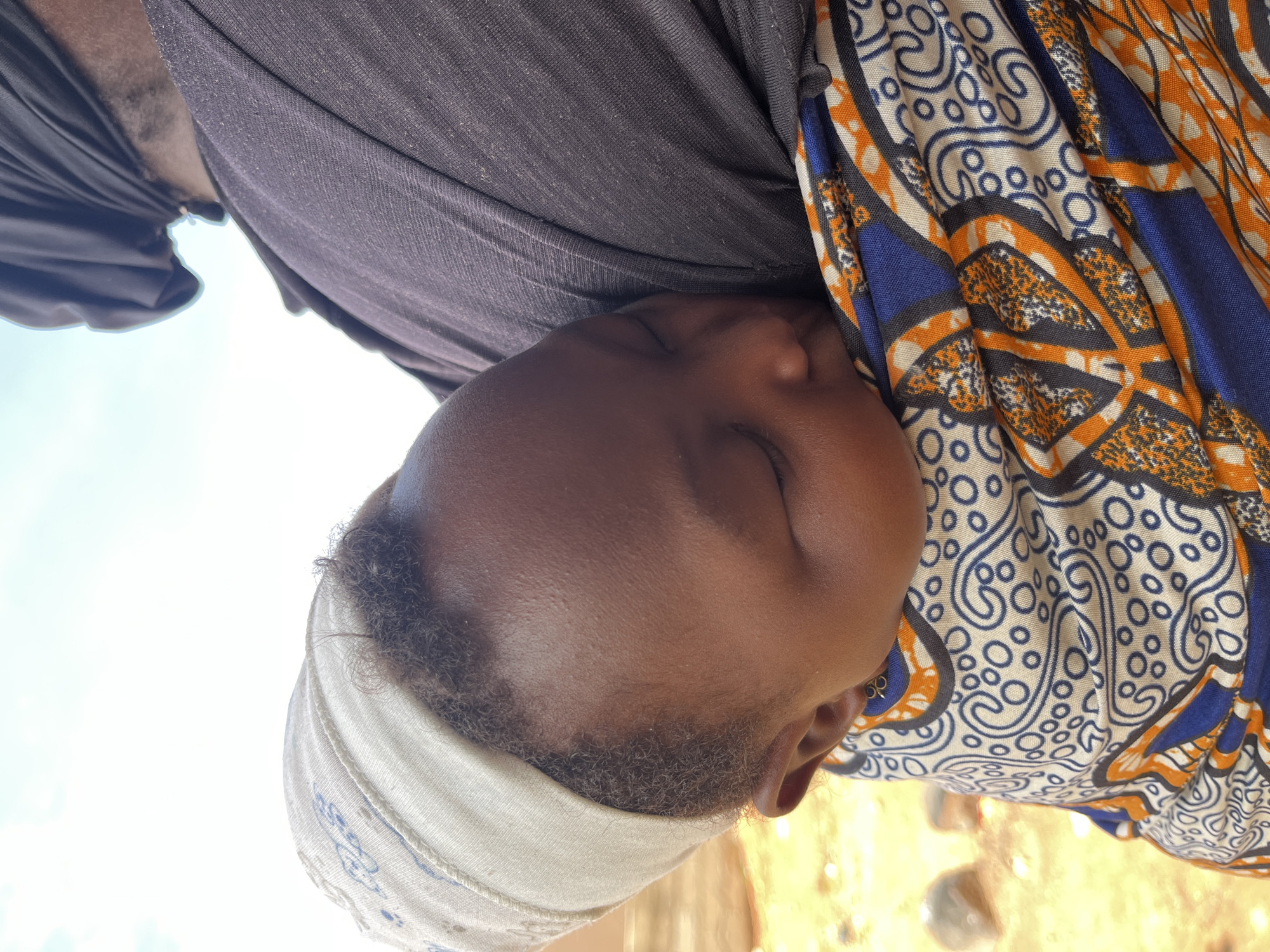 The image presents a close-up, vertical shot of a sleeping child being carried by an adult, outdoors in what is inferred to be Jos, Nigeria. The child, exhibiting dark skin, is positioned against the adult's upper back and shoulder, secured by a vividly patterned fabric wrap. The child's eyes are closed in sleep, and a light-colored, possibly off-white or beige, headwrap with faint blue patterns covers their short, curly hair. A small, simple, gold-toned earring is visible in the child's right ear. The adult, only partially visible, has dark skin and wears a dark, likely black or dark grey, short-sleeved top. A dark head covering on the adult's head is visible at the top right of the frame. The fabric wrapping the child features an intricate pattern in creamy white, royal blue, orange, and black, incorporating swirling paisley-like motifs, concentric circles, and geometric shapes, with the orange sections exhibiting a speckled texture. The background consists of a bright, light blue-white sky in the upper portion and a blurred, light brown or tan ground, possibly dirt or sand, with indistinct shapes in the lower left, indicating an unpaved outdoor environment.