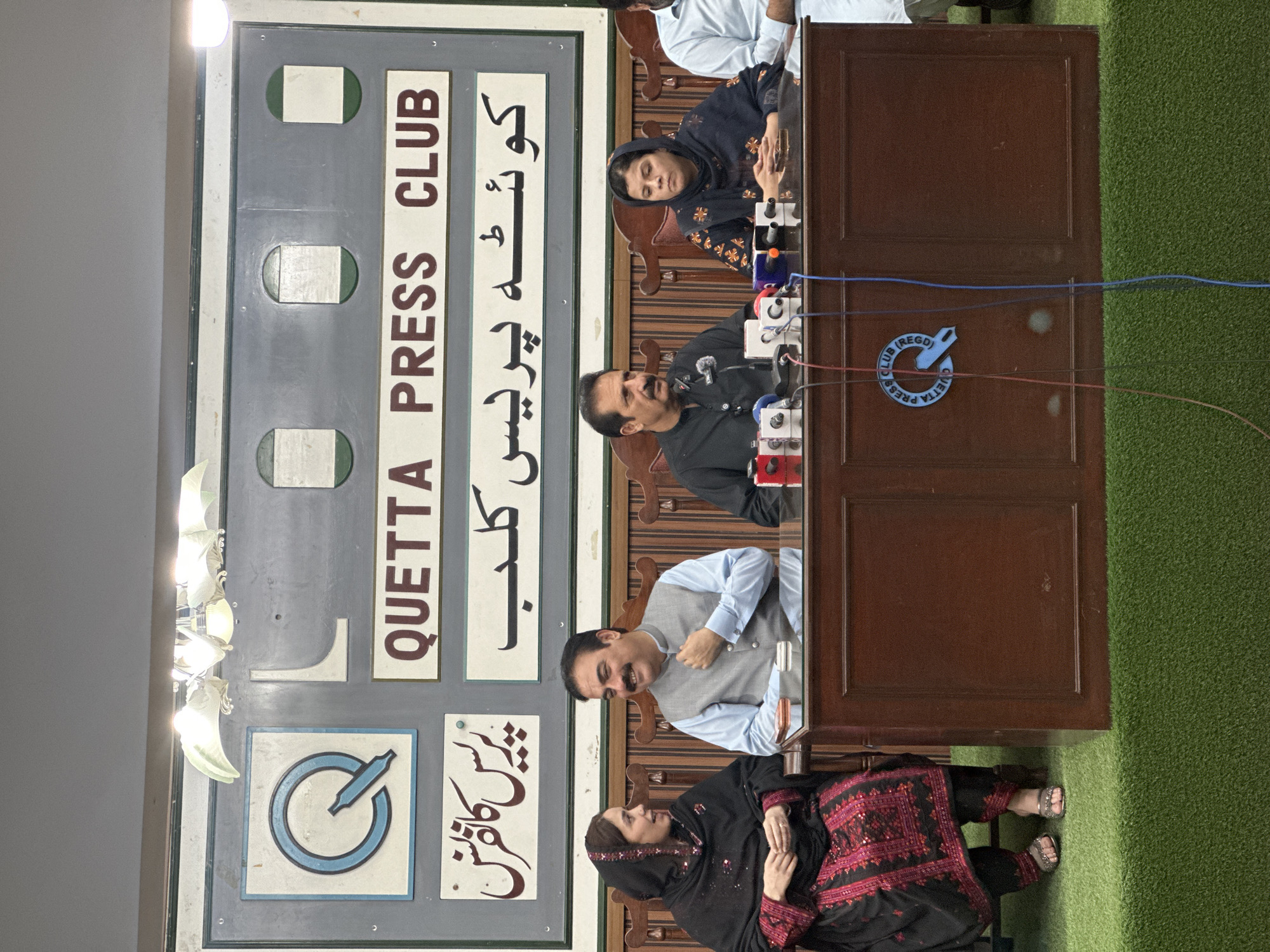 The image depicts a press conference at the Quetta Press Club, located in Quetta City Tehsil, Pakistan. Four individuals are seated behind a wooden podium. On the left, a woman wearing a black shalwar kameez with intricate red embroidery and a black hijab is seated, facing towards the right and engaged in conversation with the man next to her. The man, wearing a light blue shirt and a grey vest, is smiling and looking at the woman. To his right, another man in a black outfit with a mustache is looking forward, with a microphone in front of him. On the far right, a woman in a dark blue hijab and patterned clothing is seated, looking forward with her hands clasped. Multiple microphones are positioned on the podium, indicating a formal press event. Behind the individuals, a backdrop features the "QUETTA PRESS CLUB" logo in English and Urdu, along with other signage. The foreground shows a green carpeted platform. The lighting suggests an indoor setting.