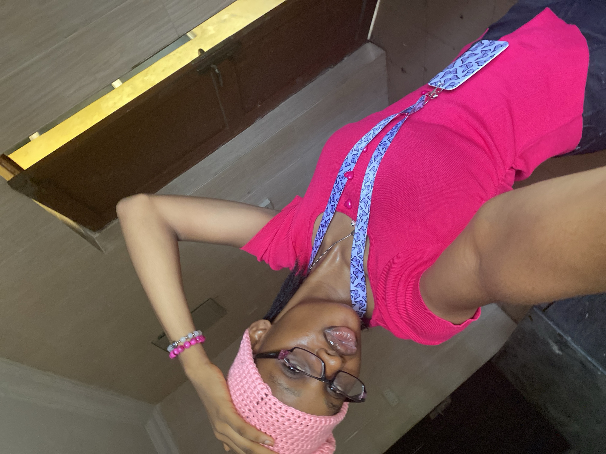 A young person with dark skin, wearing glasses and a pink knitted cap, is seen in a selfie. The person is wearing a pink top and a lanyard with a butterfly design. A pink and white beaded bracelet is visible on their left wrist. They appear to be indoors, with a brown cabinet or door visible in the background. The location is Unknown, Unknown.