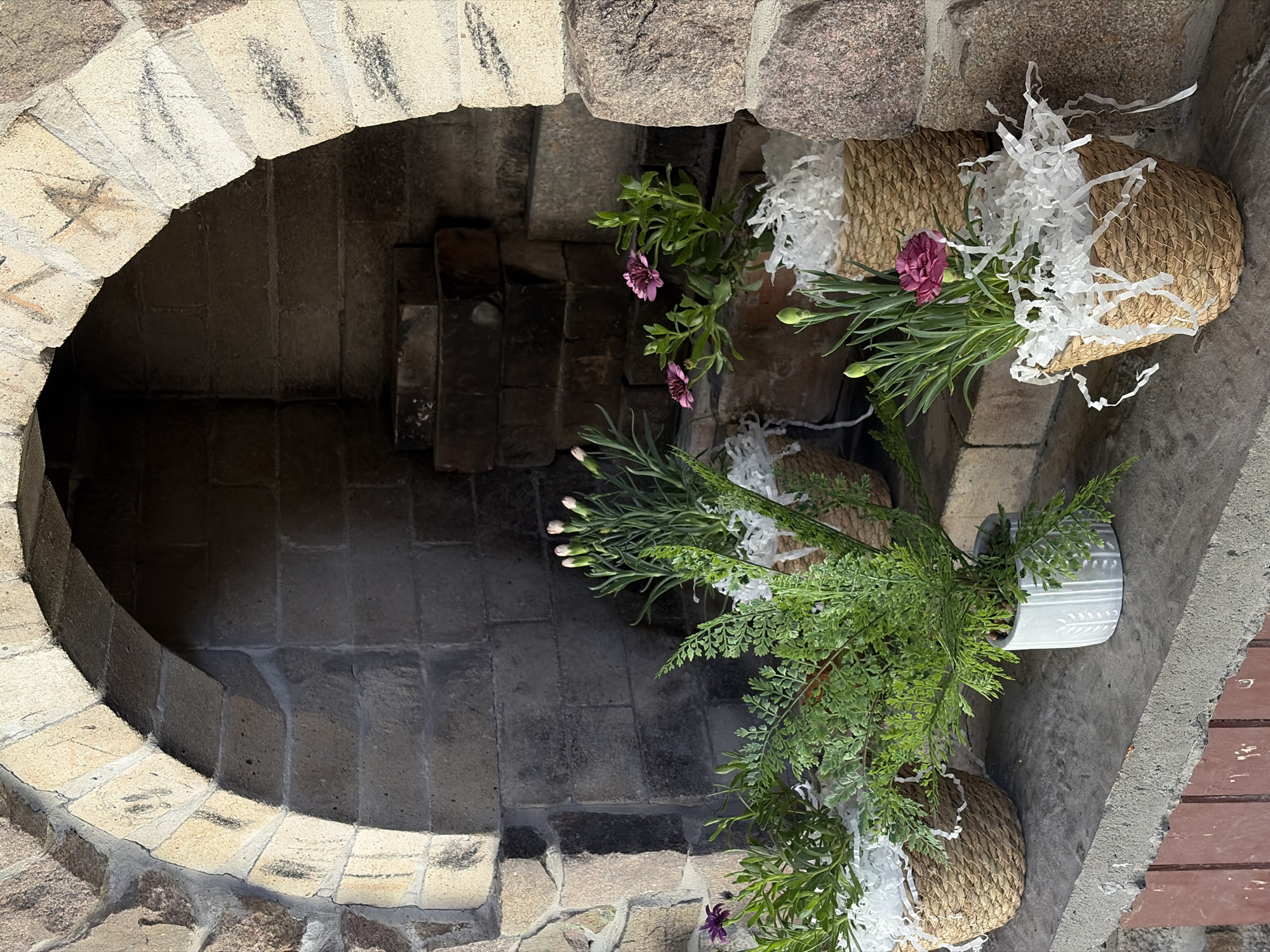 The image depicts an outdoor fireplace with a stone archway. Inside the fireplace, bricks are visible, and a shelf extends from the fireplace. On this shelf, there are four potted plants arranged in a row. The plants are in woven baskets and one white pot, filled with white shredded paper. Two of the plants have small pink flowers, one has purple flowers, and the remaining plant is a fern. The fireplace is constructed from a mix of rough-hewn and cut stone blocks. The ground in front of the fireplace appears to be concrete, with a reddish-brown wooden structure visible below the shelf. The overall scene suggests a decorative arrangement in an outdoor setting. There are no people or animals present, and no discernible Kyiv landmarks are visible.