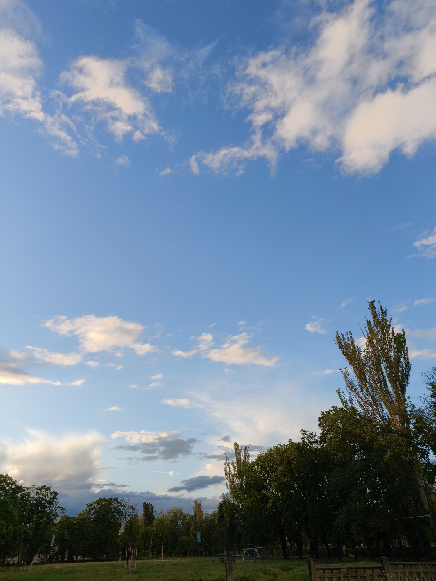 The image captures a park scene under a blue sky dotted with white clouds. In the foreground is a grassy area, with trees lining the horizon. The location is Kryvyi Rih, Ukraine. There are no people visible in the image. A wooden fence is partially visible on the right side of the image.