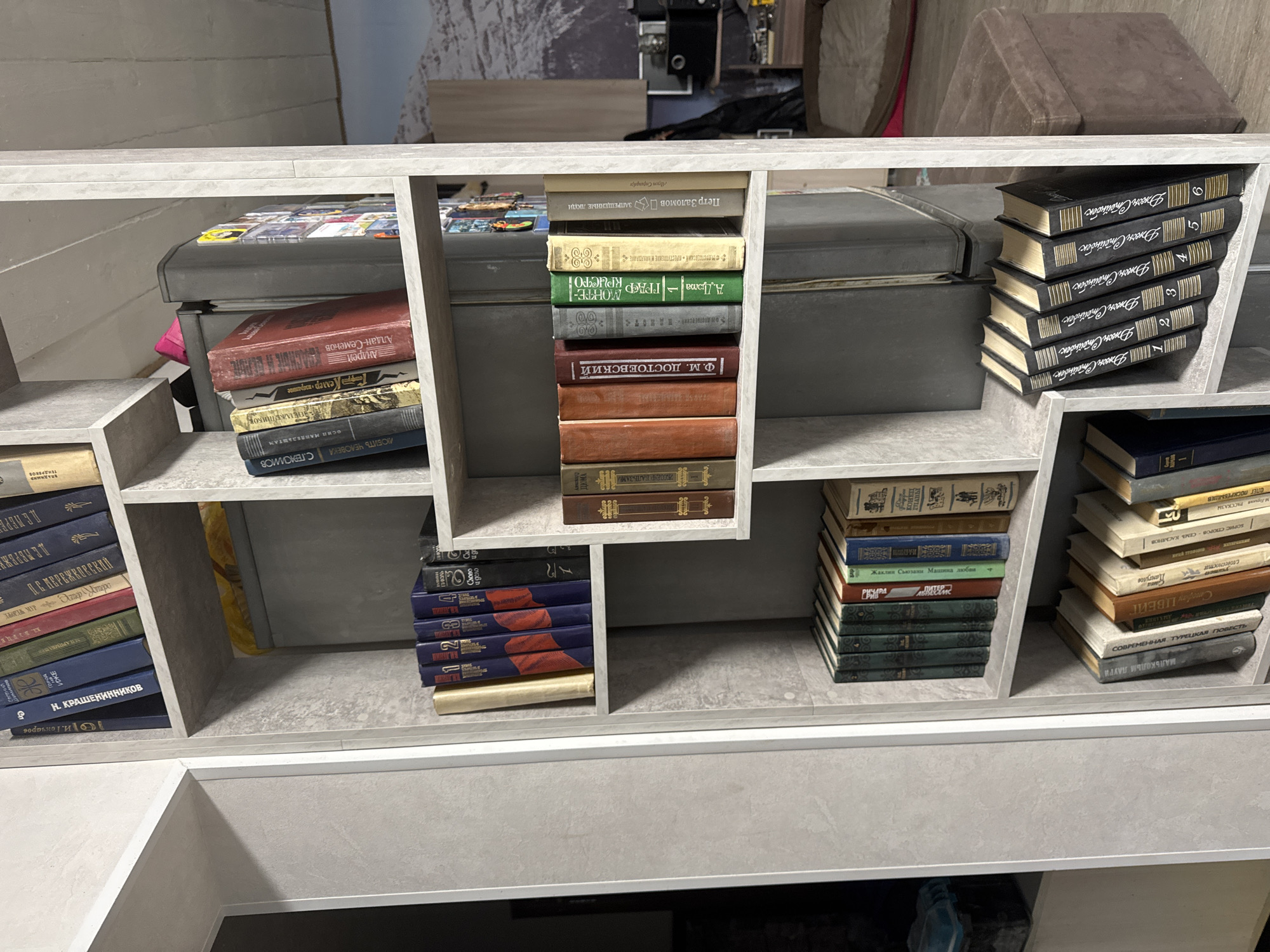 The image shows a bookshelf filled with various books. The books are stacked vertically and horizontally, and they appear to be primarily in Russian. The bookshelf is made of a light grey material and has a geometric design with open shelves. There are no people visible in the image. The room's decor suggests an indoor environment in a residential or office space. The spines of the books contain legible text, including the names of authors and titles.