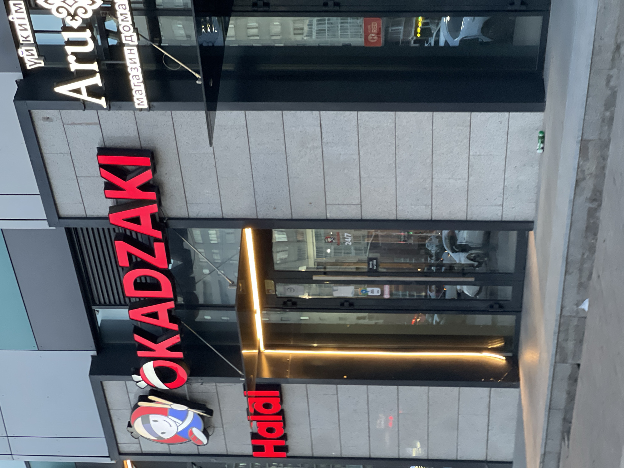 The image shows the exterior of a business establishment in Astana, Kazakhstan. The facade features stone cladding and glass doors. A prominent red neon sign reads "OKADZAKI" with a stylized cartoon character to its left, holding chopsticks. Below this, another red neon sign says "Halal". To the right of "OKADZAKI," a white neon sign displays "Aru," with Russian text above and below it. Reflections in the glass doors reveal vehicles and buildings. The ground in front of the establishment is paved, with a slight incline, and there is some litter visible on the ground. The lighting suggests it is either early morning or late evening.