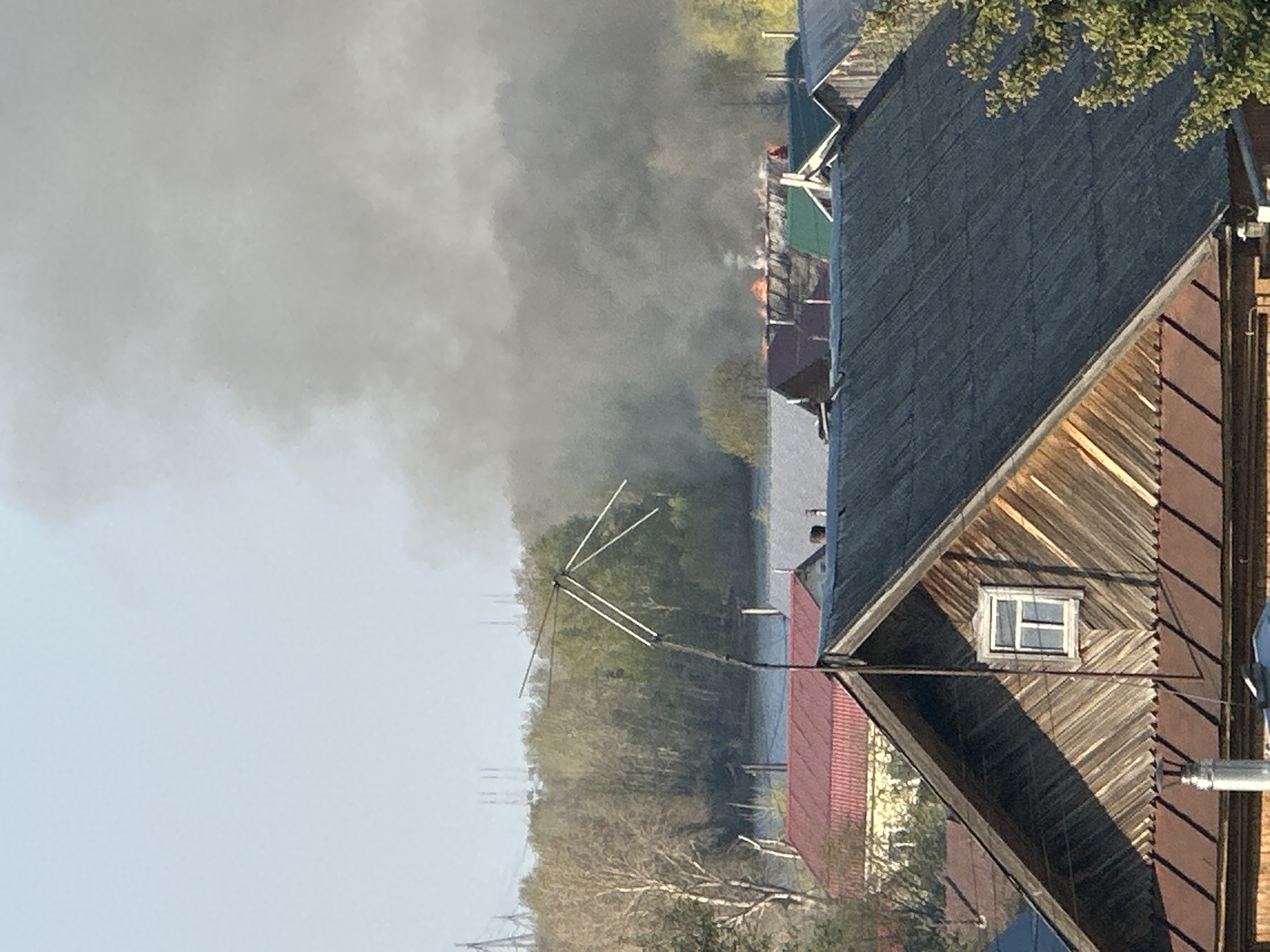 The image shows a fire in a residential area, likely in the city of Chaikovsky, Russia. A large plume of dark smoke rises in the background, indicating a substantial fire. Flames are visible amidst the smoke, suggesting active burning. In the foreground, there is a wooden house with a gabled roof and a small window. The siding of the house is a mix of light and dark wood, indicating aging or possibly fire damage. An antenna is attached to the side of the house. Other houses and rooftops are visible in the distance, along with trees, possibly bordering a body of water. There are also power lines in the distance. The scene suggests a serious fire event impacting the residential neighborhood.