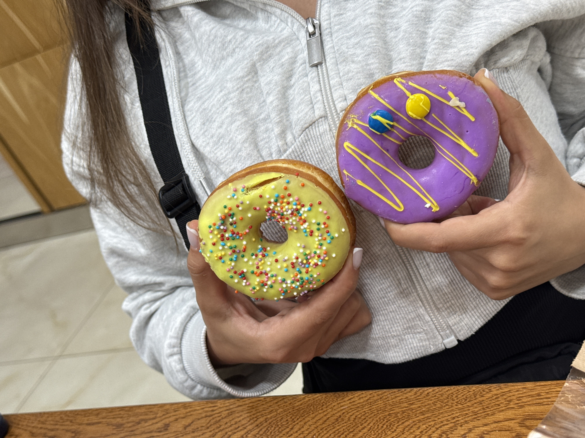 A person holds two donuts in their hands. The donut on the left has yellow frosting and multi-colored sprinkles. The donut on the right has purple frosting, yellow stripes, and three candy decorations. The person wears a grey zip-up hoodie and a black strap is visible over their left shoulder. The image appears to be taken indoors, with a wooden table in the foreground and a tan wall in the background. The location is not evident in the image.