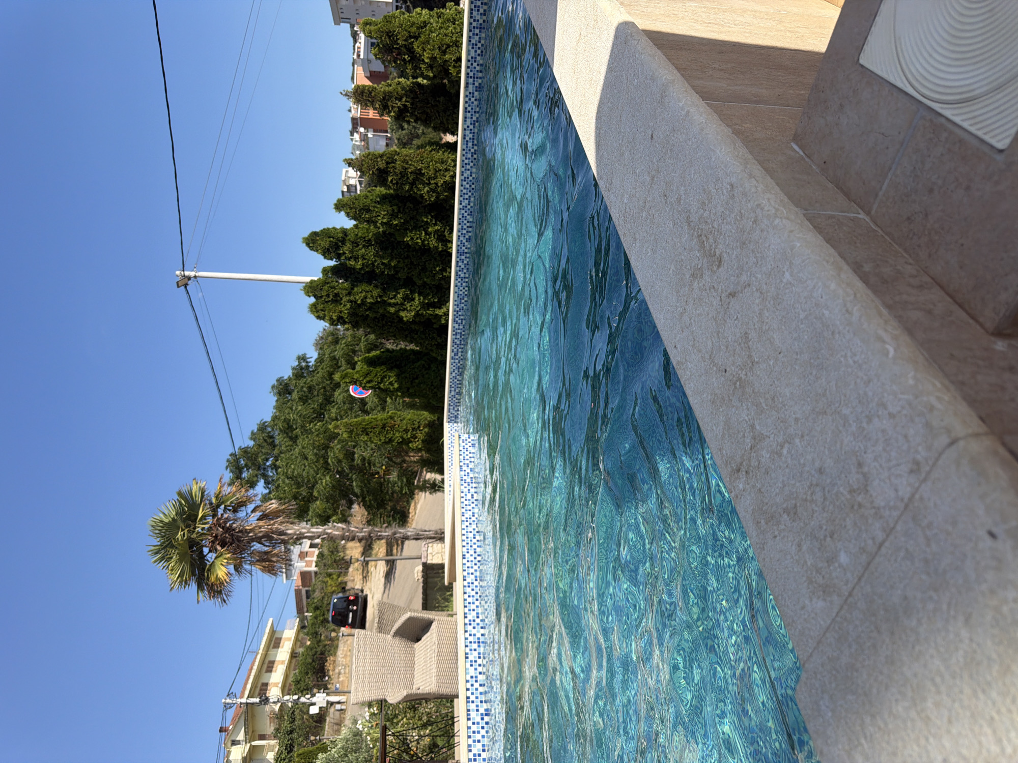 The image shows a bright, sunny day in Pečurice, Montenegro. A swimming pool with clear blue water is in the foreground, with a tiled edge. Beyond the pool, there are cypress trees, a palm tree, and houses on a hillside under a clear blue sky. A black van is parked on a road in the background. A wicker chair is visible next to the pool.