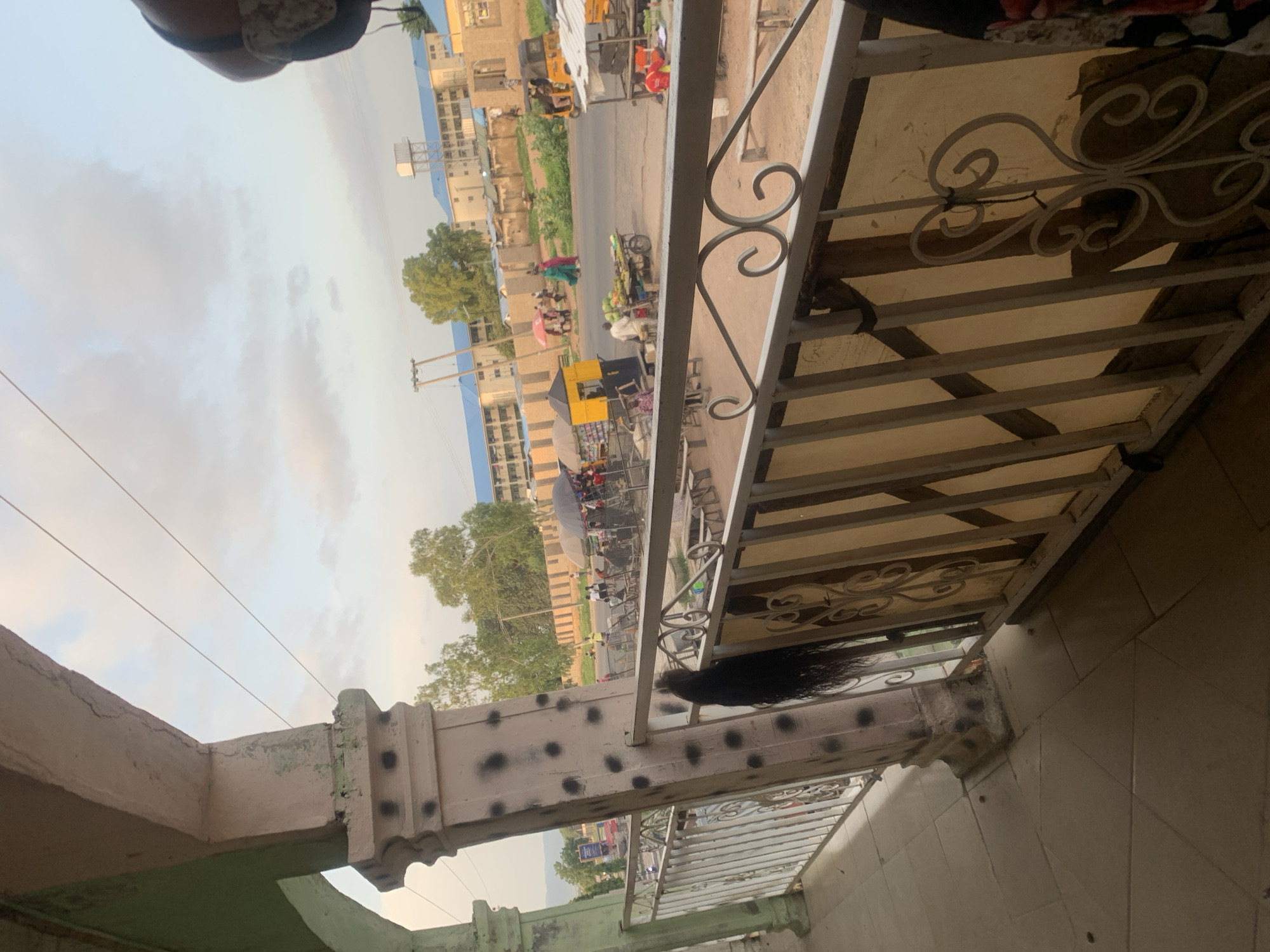 A view from a balcony looks out onto a street scene in Jalingo, Nigeria. In the foreground, a decorative metal railing with a beige-colored backing separates the balcony from the street. A pale pink column with black circular markings stands to the left, partially obscuring the view. In the middle ground, a busy street is visible with various market stalls and vehicles. People are seen walking and interacting at the stalls. A yellow and blue kiosk is situated on the left side of the street, and further down, a man is selling produce from a cart. A tricycle and a tuk-tuk are also present on the road. In the background, there are buildings with blue roofs and a water tower. The sky is partly cloudy.