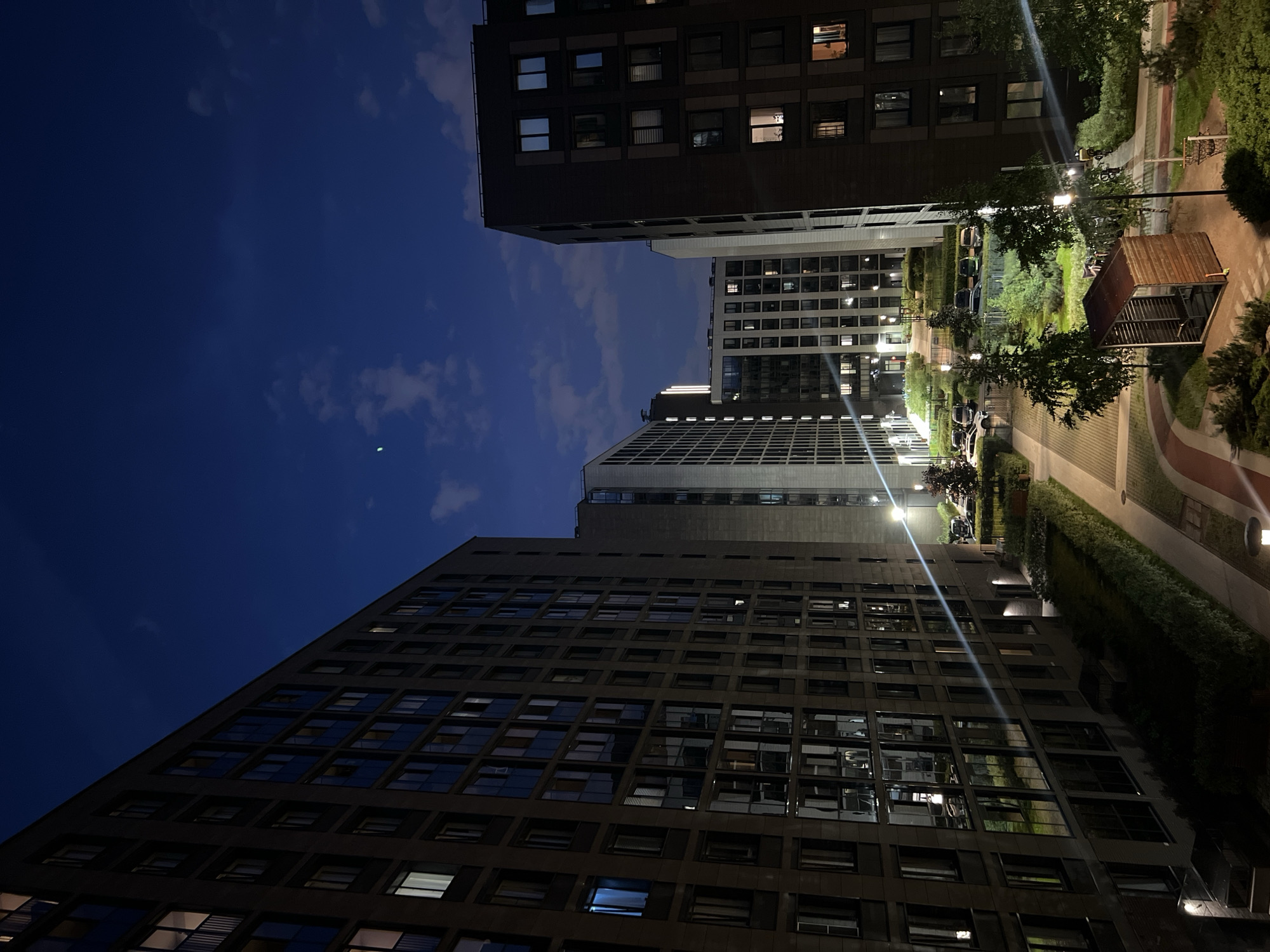 This is a nighttime, eye-level, outdoor, medium shot of a courtyard between several modern residential buildings in the city of Unknown, Russia. The sky is a deep blue with scattered, wispy clouds and a small green light, possibly an aircraft or satellite, is visible in the upper portion of the sky. The buildings are multi-story and appear to be apartment complexes, with many windows lit from within, suggesting occupancy. The architecture is contemporary, with clean lines and dark facades.
The courtyard area features paved walkways, grassy areas, and trees. A small, wooden gazebo or shelter is situated in the foreground, and a few parked cars are visible in the background. Streetlights illuminate various parts of the courtyard, casting long shadows and highlighting the architectural details of the buildings. The scene conveys a sense of urban residential tranquility at night. There are no people visible in the image.