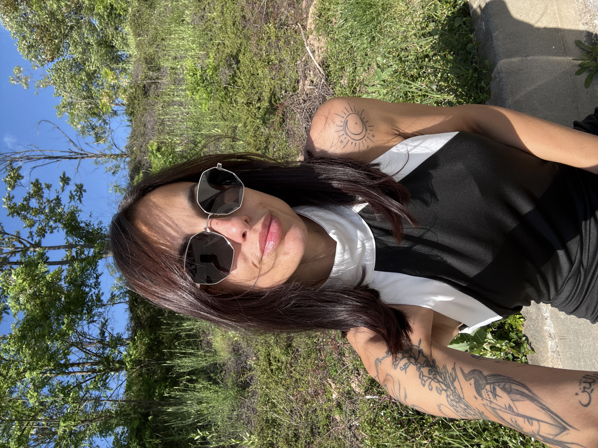 A woman with long, dark brown hair is taking a selfie outdoors. She is wearing sunglasses and a black and white top. She has tattoos on her arms and shoulder. The background shows green grass, trees, and a blue sky. A concrete surface is visible in the background, possibly a sidewalk or ledge. The location in the city of Pleven, Bulgaria cannot be determined from the image.