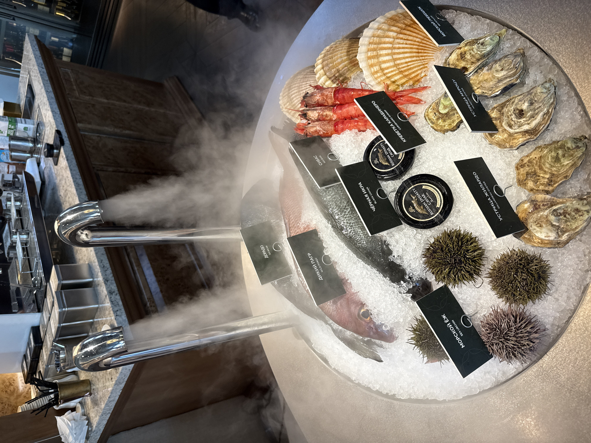The image depicts a seafood display at what appears to be a restaurant or market in Moscow, Russia. In the foreground, a circular arrangement of fresh seafood rests on a bed of crushed ice. The seafood includes several types of fish, including what is labeled as "Dorado" and "Wild Porgy," as well as a specimen identified as "Sea Bass." There are also several bright red Carabinero shrimp, some with their heads intact and tails exposed. Several large scallop shells are also present, adding to the visual appeal. The display is enhanced by a dramatic effect of steam or mist emanating from two chrome taps positioned above the ice.
Surrounding the main seafood items are smaller delicacies. Two jars of "Russian Dynasty Caviar" are visible, along with several sea urchins, each with spiky exteriors, and a collection of oysters, some still in their shells. Black labels with white text are strategically placed to identify each type of seafood.
In the background, a counter area is visible with various items suggestive of a food service establishment. Stacked white paper cups, black utensils in a holder, and metal containers are arranged on a speckled countertop. Beyond this, a commercial coffee machine or similar equipment is partially visible, and wooden cabinetry lines the lower portion of the background. The overall lighting suggests an indoor setting, likely with ambient restaurant lighting. No people are clearly visible in the frame, though the presence of a person's leg and shoe in the lower right corner suggests at least one individual is present off-camera.