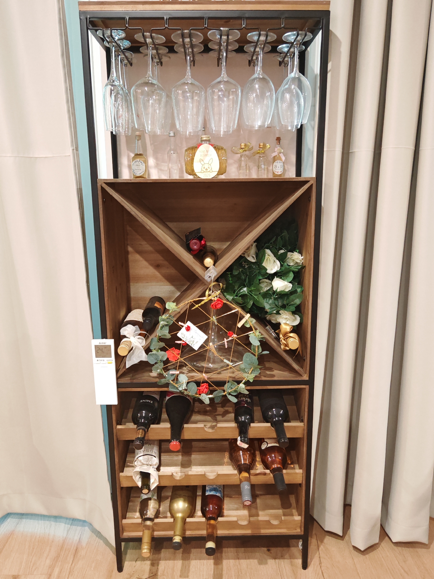 The image shows a wooden wine rack with several bottles of wine and glassware. The wine rack is tall and narrow, with multiple shelves. The top shelf has a rack for hanging wine glasses. There are also some decorative items on the shelves, including a wreath with red flowers and a bouquet of white roses. The wine rack is placed in front of a cream-colored curtain. The floor is made of light-colored wood. A white remote control with a display showing "24°C" is visible on the left side of the wine rack. The scene appears to be an indoor setting, possibly a home or a store. No people are visible in the image. There is no information in the image to suggest a location in Plovdiv, Bulgaria.