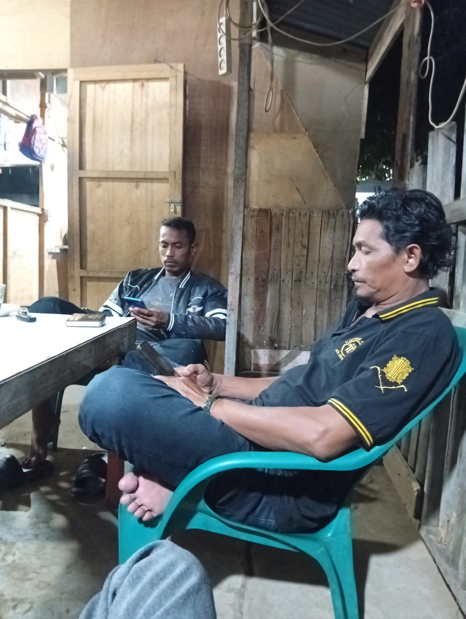 In Lhokseumawe, Indonesia, two adult males are observed seated in a rustic, wood-paneled interior, suggestive of a dwelling or informal gathering area. A third individual's legs, clad in grey fabric, are partially visible in the immediate lower foreground.
The man in the right foreground, exhibiting dark, curly hair and a beard, is seated in a teal plastic chair. He wears a black polo shirt with yellow stripes on the collar and sleeves, and two yellow embroidered emblems on the left chest. He is dressed in dark blue jeans and is barefoot. His right foot rests on his left thigh, and he holds a black smartphone with both hands, directing his gaze downwards towards the screen. A green wristband is present on his left wrist.
In the midground left, a second man with short dark hair and facial stubble is seated behind a rectangular wooden table with a light-colored top. He wears a dark bomber jacket featuring white stripes on the sleeves, layered over a grey t-shirt, and dark pants. He holds a light blue smartphone in his right hand, also looking downwards at the screen. On the table, to his left, are a small, dark-colored book and a black remote control. A pair of black slip-on sandals are visible on the light grey concrete floor beneath the table, near his feet.
The room's walls are constructed from light-toned wooden planks and vertical beams. Exposed white electrical wiring runs along the walls, and a white three-pin electrical outlet is mounted on the upper right wall. A colorful, small bag is hanging on a structure to the far left. Dark foliage is partially visible through a gap in the wooden structure on the far right, suggesting an exterior beyond. The ambient lighting appears dim, consistent with an indoor evening or dimly lit setting.