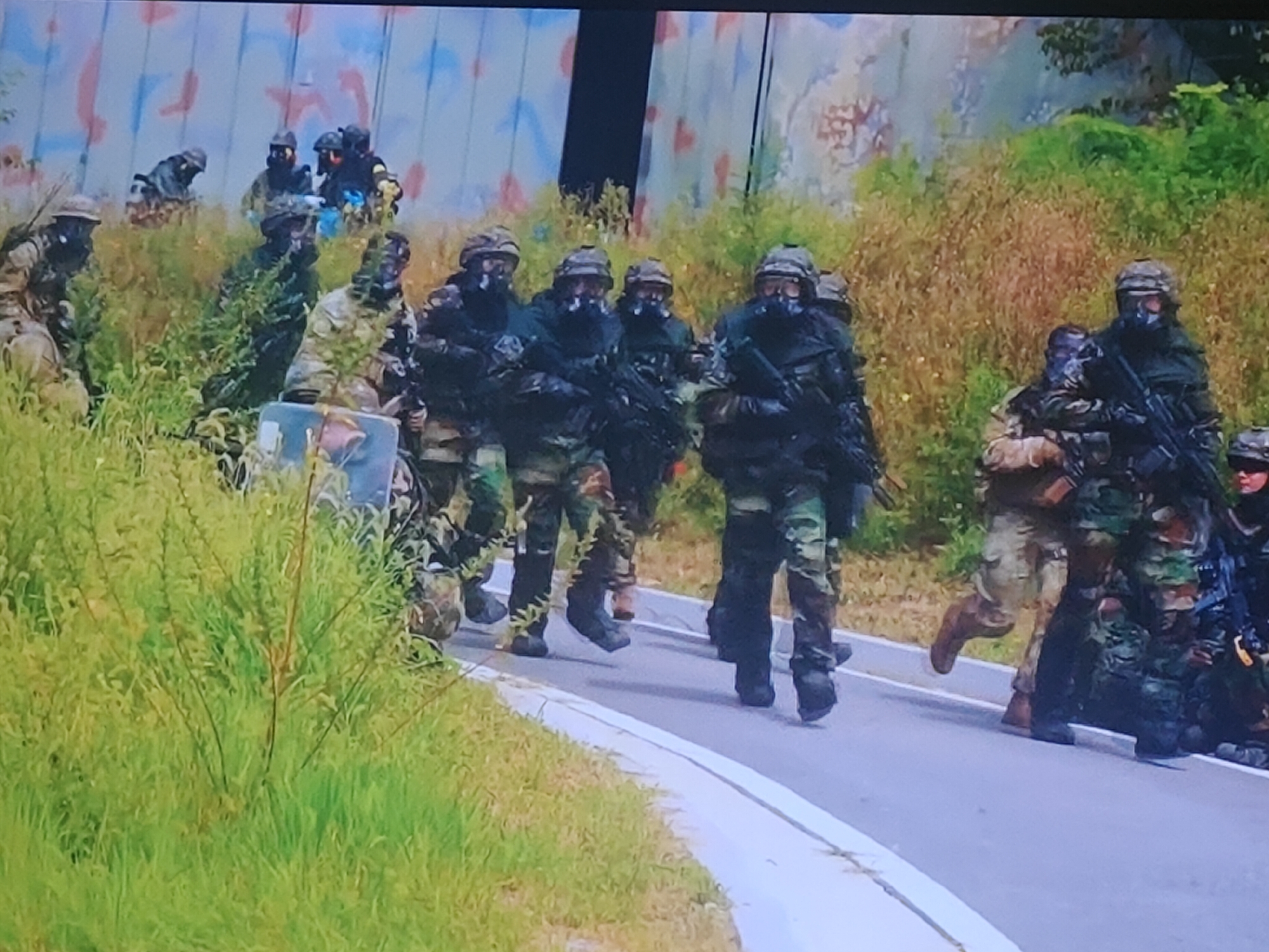 In a suburban setting, possibly in Molenbeek-Saint-Jean - Sint-Jans-Molenbeek, Belgium, a group of individuals in tactical gear, including helmets and gas masks, are participating in what appears to be a training exercise or simulation. They are dressed in camouflage uniforms and carry firearms, suggesting a scenario involving a security or military operation. The environment features a wall with a red and blue abstract pattern in the background, overgrown vegetation, and a paved road with a white line. The individuals are positioned on the road and in the grassy area beside it, some walking and others standing, all appearing to be focused and ready. One individual in the foreground is holding a transparent shield, adding to the impression of a staged event.