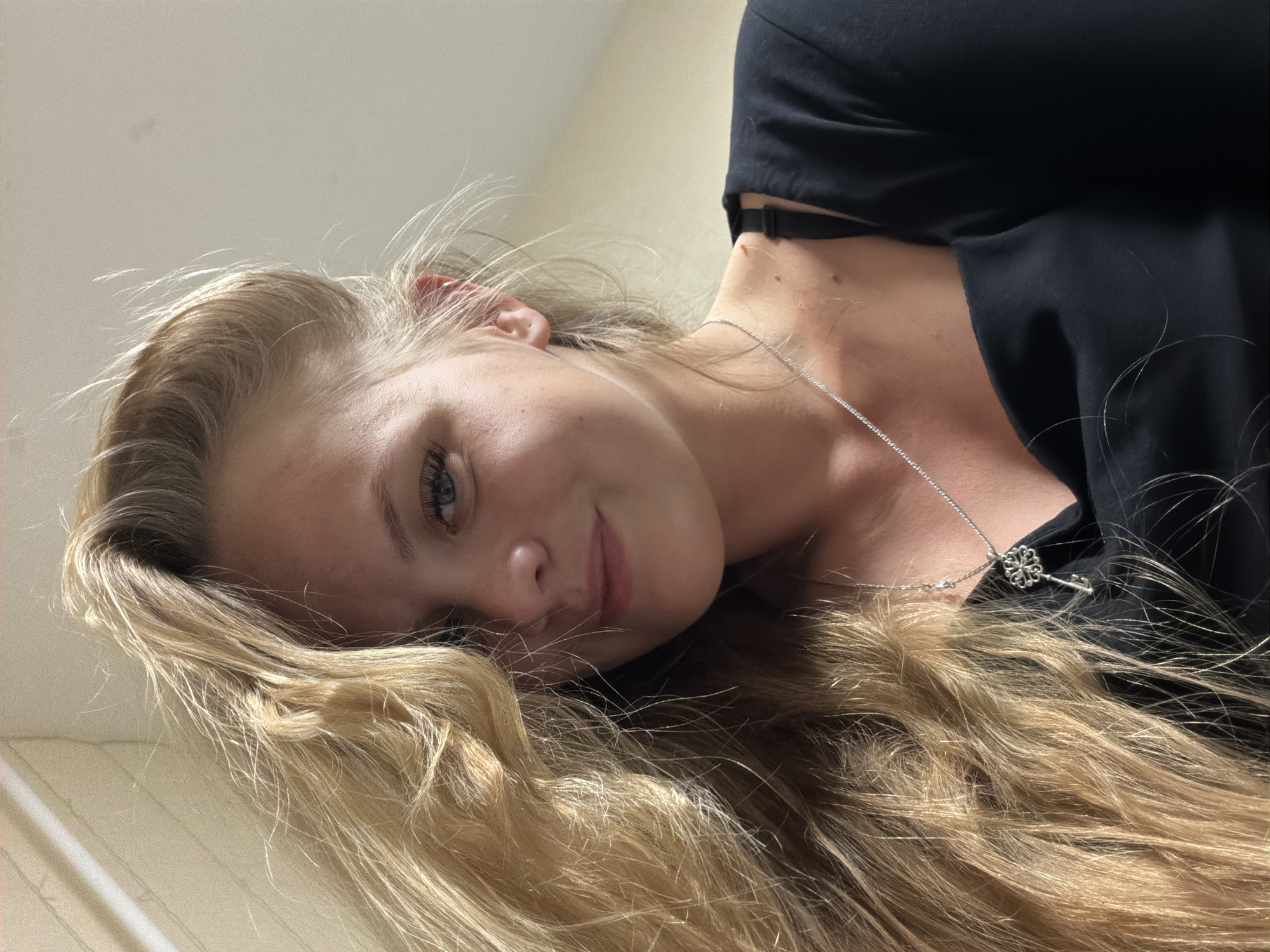 The image is a close-up, low-angle selfie of a young woman with long, blonde hair and blue eyes. She is wearing a black top and a silver necklace with a key pendant. The background is a plain wall with some indistinct lighting. There are no discernible features that would indicate a specific location in Izhevsk, Russia. The image captures a moment of casual self-expression, with the woman's hair falling across her face and a slight smile on her lips. The lighting suggests an indoor setting, possibly a home.