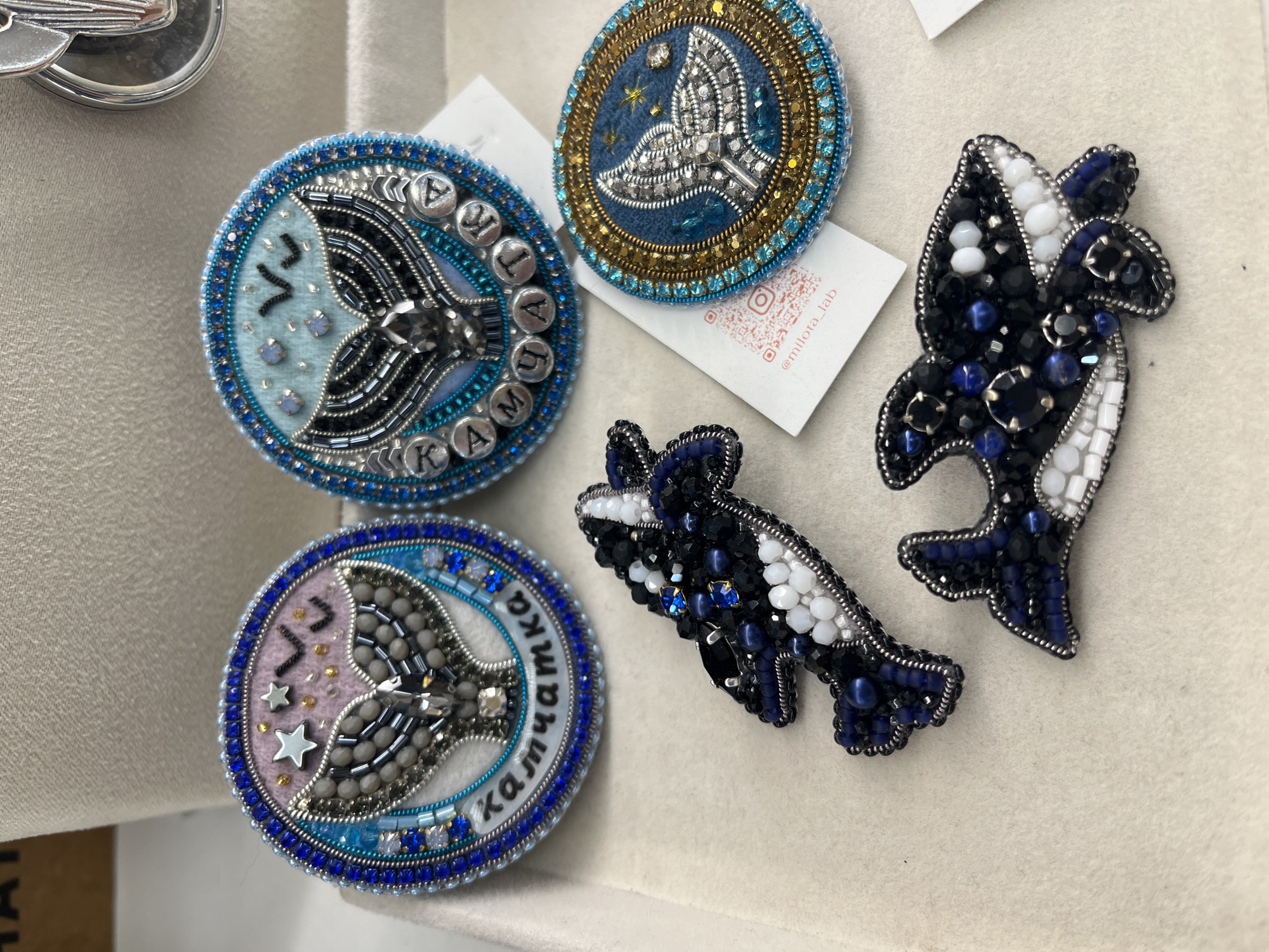 The image displays a collection of beaded brooches, likely a product display. In the upper left, two circular brooches are visible. The left one features a light purple background with a silver whale tail made of beads, stars, and what appear to be small black bird silhouettes. The text "камчатка" is embroidered in a circular pattern around the whale's tail. The right circular brooch has a light blue background with a similar beaded whale tail, and what seem to be small light blue rhinestones and black bird silhouettes. The text around this brooch is also "камчатка". To the right and slightly lower, a third circular brooch features a dark blue background with a silver beaded whale tail, embellished with gold and blue beads along its border. Below these circular brooches are two more brooches shaped like orcas (killer whales). The front orca brooch is predominantly black and white, with blue and clear bead accents. The second orca brooch, partially visible behind the first, shares a similar color scheme and beading style. The brooches are laid out on a neutral-colored, textured surface, possibly a display cloth or a shelf. In the background, a portion of a tan, textured surface is visible, possibly part of a booth or display structure. A white label with a QR code and "@milota_lab" printed on it is partially visible beneath the circular brooches. The overall scene suggests a retail or craft fair setting. There are no people visible in the image. The location of Светлый, Russia is not ascertainable from the image itself.