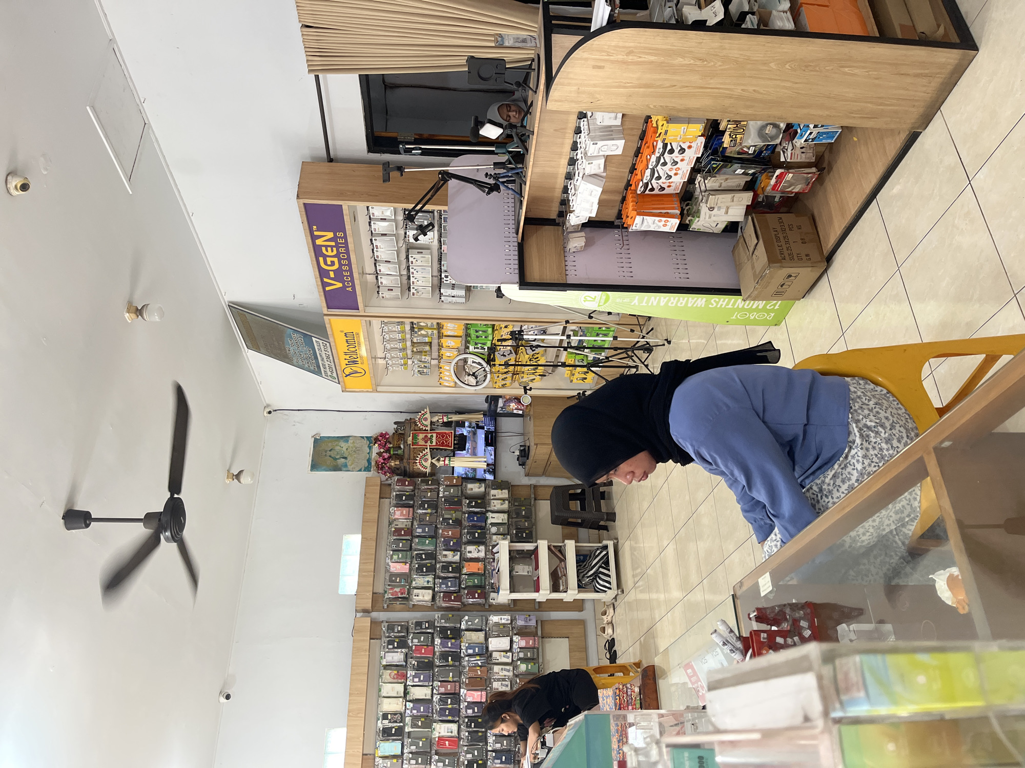 The image depicts the interior of a retail store located in North Kuta, Indonesia, specializing in mobile accessories. A ceiling fan is visible overhead, along with several security cameras mounted on the white ceiling. The store is organized with display shelves showcasing numerous mobile phone cases, chargers, headphones, and other accessories. Two women are present in the store. One woman, wearing a black hijab and blue jacket, is seated at a counter and appears to be working or attending to customers. Another woman, also wearing a black hijab and black top, is standing and looking towards the shelves. The store features a tiled floor, wooden display units, and a television screen displaying content. Artificial plants and decorative items are also visible in the background. A stack of boxes is placed on the floor near a display shelf. The overall atmosphere suggests a typical small retail establishment.