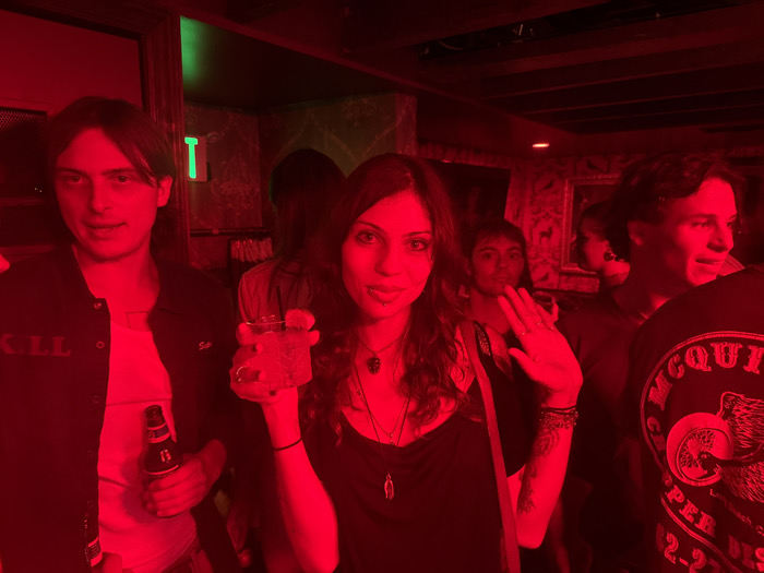 The image depicts an indoor scene in a crowded social venue within West Hollywood, United States of America, illuminated primarily by a strong red light. Five individuals are visible. On the left, a male subject with medium-length dark hair, wearing a dark jacket over a lighter shirt, holds a brown glass bottle in his right hand, looking towards the camera. To his right, a central female subject with long dark hair, wearing a dark top with visible necklaces, holds a clear glass containing a beverage in her raised left hand. Her right forearm, bearing tattoos, is raised with the palm facing forward. She is looking at the camera with a slight smile. Behind and to the right of the central female, a person with dark hair is partially visible. Further right, another person with dark hair is seen looking towards the camera. On the far right, a male subject, partially viewed from the back-right, wears a dark shirt with a prominent circular graphic featuring text, including "MCQUIRE," and an animal emblem. The background reveals dark, possibly wooden, paneling on the walls and ceiling, with a distinct vertical green light source positioned in the upper-left quadrant of the frame.