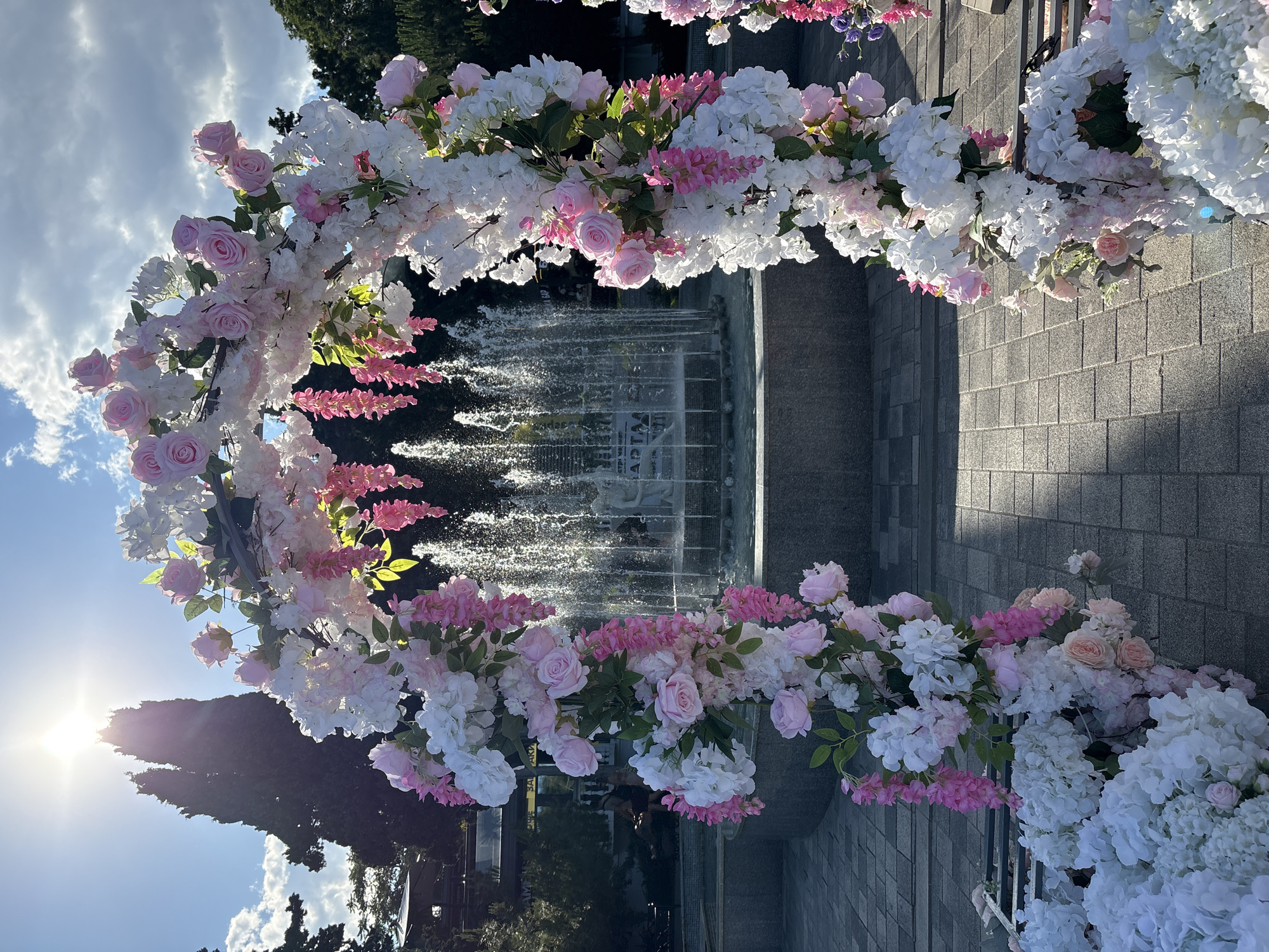 This image depicts a floral archway adorned with white and pink artificial flowers, positioned in front of a public fountain in Alushta, Ukraine. The sun is shining brightly, casting long shadows on the paved ground. The fountain is active, with water spraying upwards. In the background, there are trees and some buildings. No people are clearly visible interacting with the archway or the fountain. The scene appears to be set up for an event or as a decorative feature in an outdoor public space.