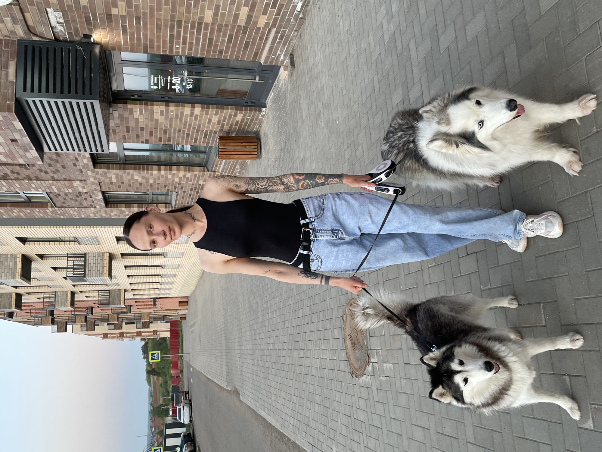 A person with tattoos, wearing a black tank top and blue jeans, holds leashes attached to two Siberian Huskies. The dogs are lying on a paved sidewalk. The person is standing in front of a brick building with a storefront showing operating hours. A street with a crosswalk sign is visible. The city of Izhevsk, Russia, is identifiable from the architecture, the crosswalk sign, and the license plate on the vehicles in the background.
