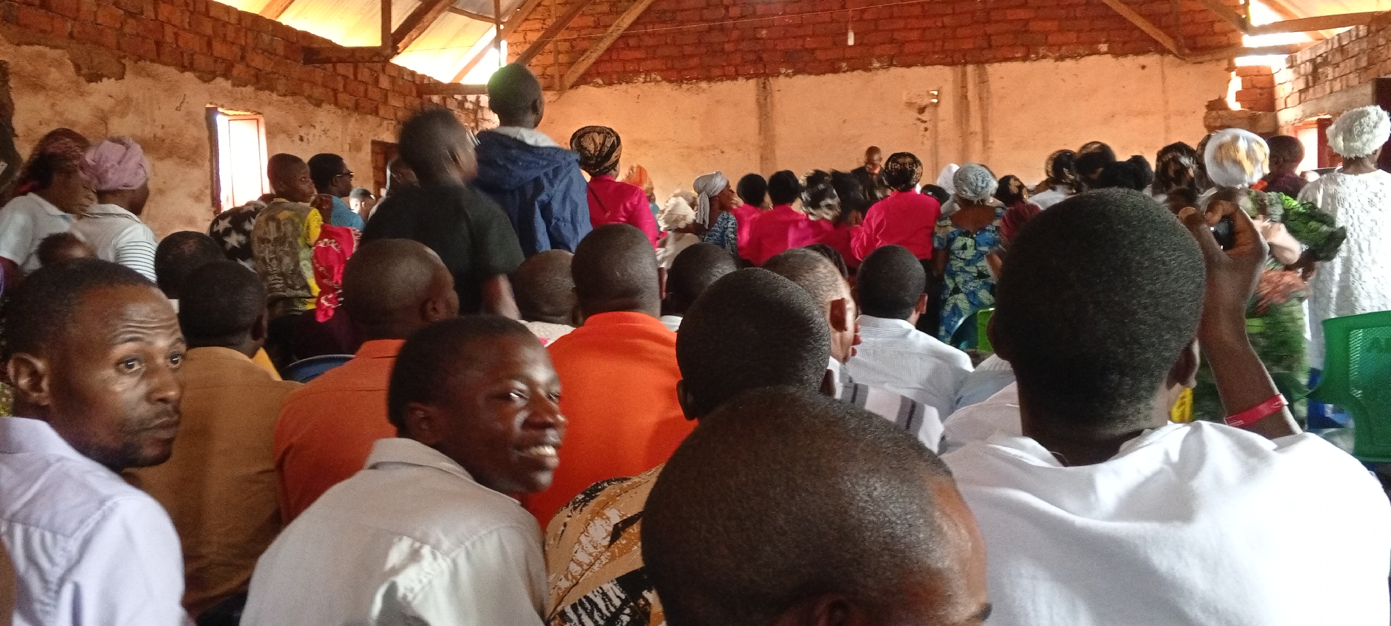 A medium, slightly high-angle shot depicts a large indoor gathering in Ngalula, Democratic Republic of the Congo. The location appears to be a meeting hall or simple church, with unfinished brick walls and a metal roof supported by wooden beams. The room is brightly lit by natural light entering through windows.
The focus is on the audience, predominantly Black, seated closely together. Most are facing forward, presumably toward a speaker or activity. Men are seen in shirts of various colors, including white, orange, light blue, and brown. Women are wearing a variety of colorful fabrics. Some people in the foreground are closer to the camera. The people in the middle and background are standing. Some are wearing head coverings. There are a few children present in the crowd. One person in the foreground has raised their arms, possibly recording with a phone. A green plastic chair is visible on the right side of the frame.