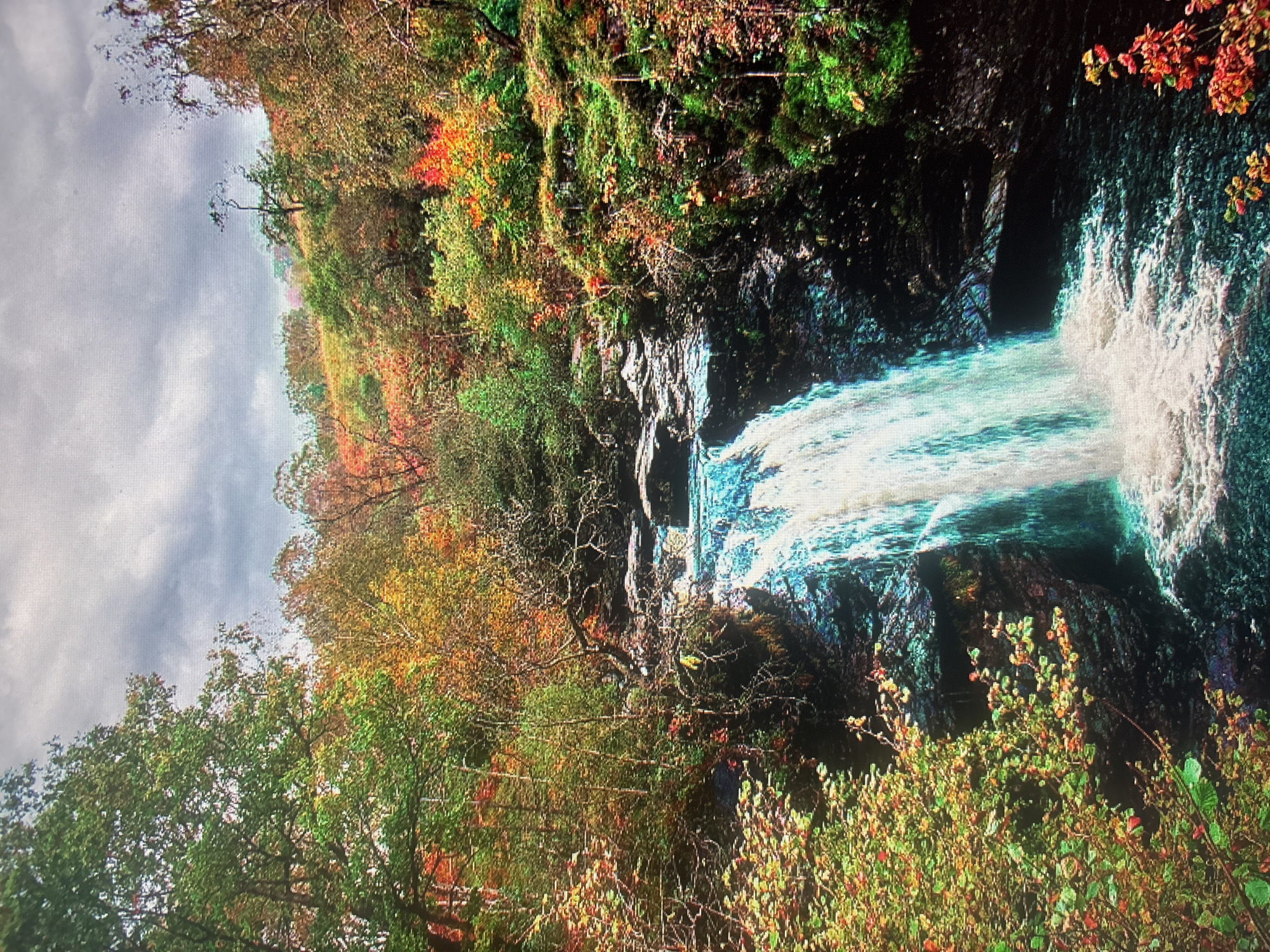 The image depicts a waterfall cascading down a rocky cliff amidst an autumnal forest. The trees exhibit a range of colors from green to yellow to red. The sky is overcast. The waterfall appears to be located in a wooded area. There is no indication of a city or urban environment. There are no people visible in the image. The location in the city of Durmersheim, Germany cannot be determined from the visual information.