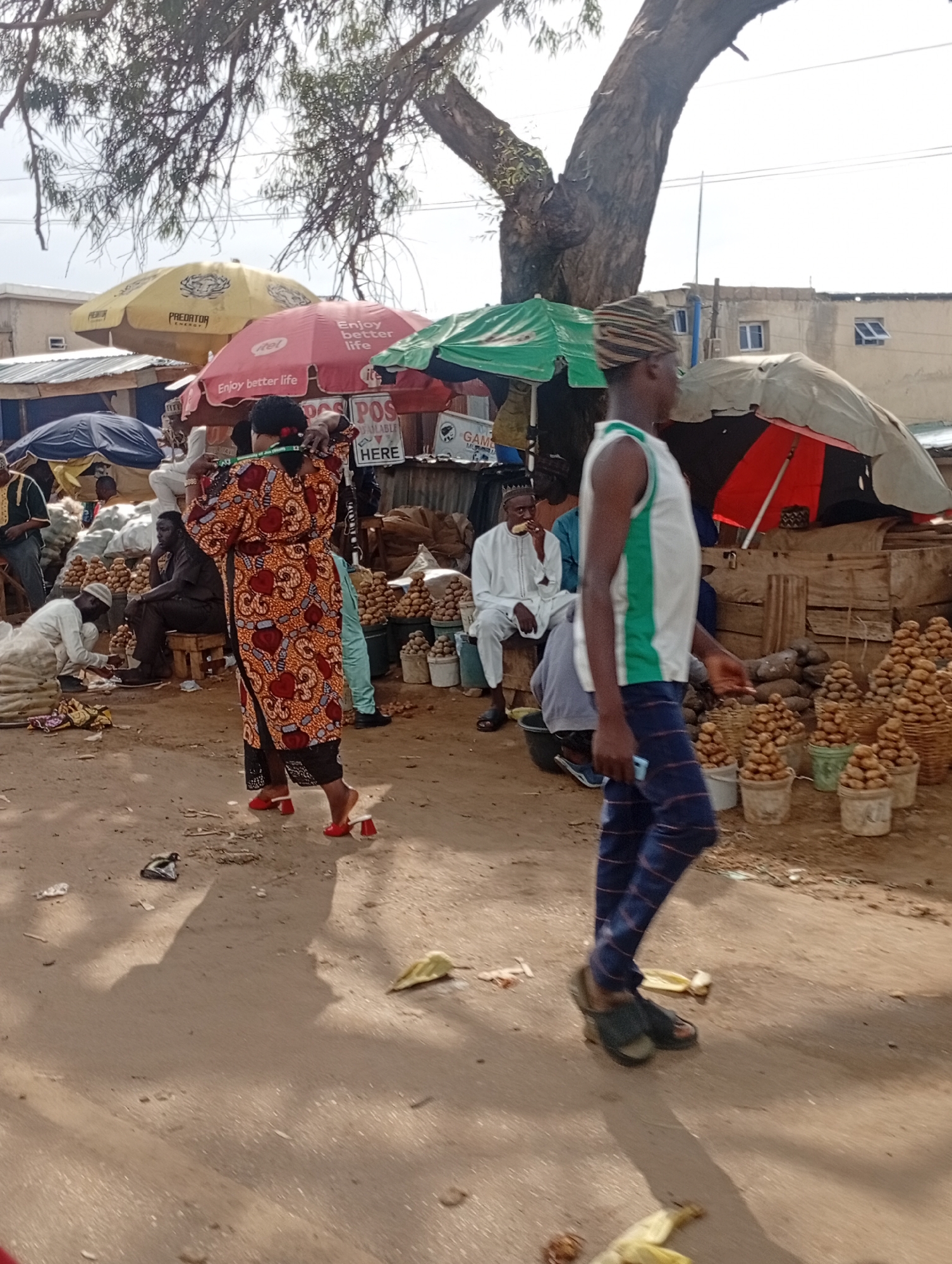 In Rafin Par, Nigeria, a busy outdoor market scene unfolds under the shade of a large tree. Several individuals are present, some tending to goods and others moving through the marketplace. A person in the foreground, facing right, is wearing a white tank top with green trim and patterned blue trousers. They are walking and holding a light-colored object, possibly a phone, in their right hand. To their left, a woman in a vibrant, patterned orange and red dress with black pants is standing and appears to be measuring something with a tape measure.
The market is filled with various stalls and goods. Several large umbrellas, including a yellow "PREDATOR ENERGY" umbrella and a pink umbrella with the "itel" logo and the phrase "Enjoy better life," provide shade. Stacks of potatoes are visible in baskets and buckets, indicating produce sales. Other market elements include wooden crates, sacks of what appear to be produce, and general debris on the dirt ground. In the background, buildings are visible, suggesting the market is situated within a populated area of the city. The overall atmosphere is one of active commerce and daily life.