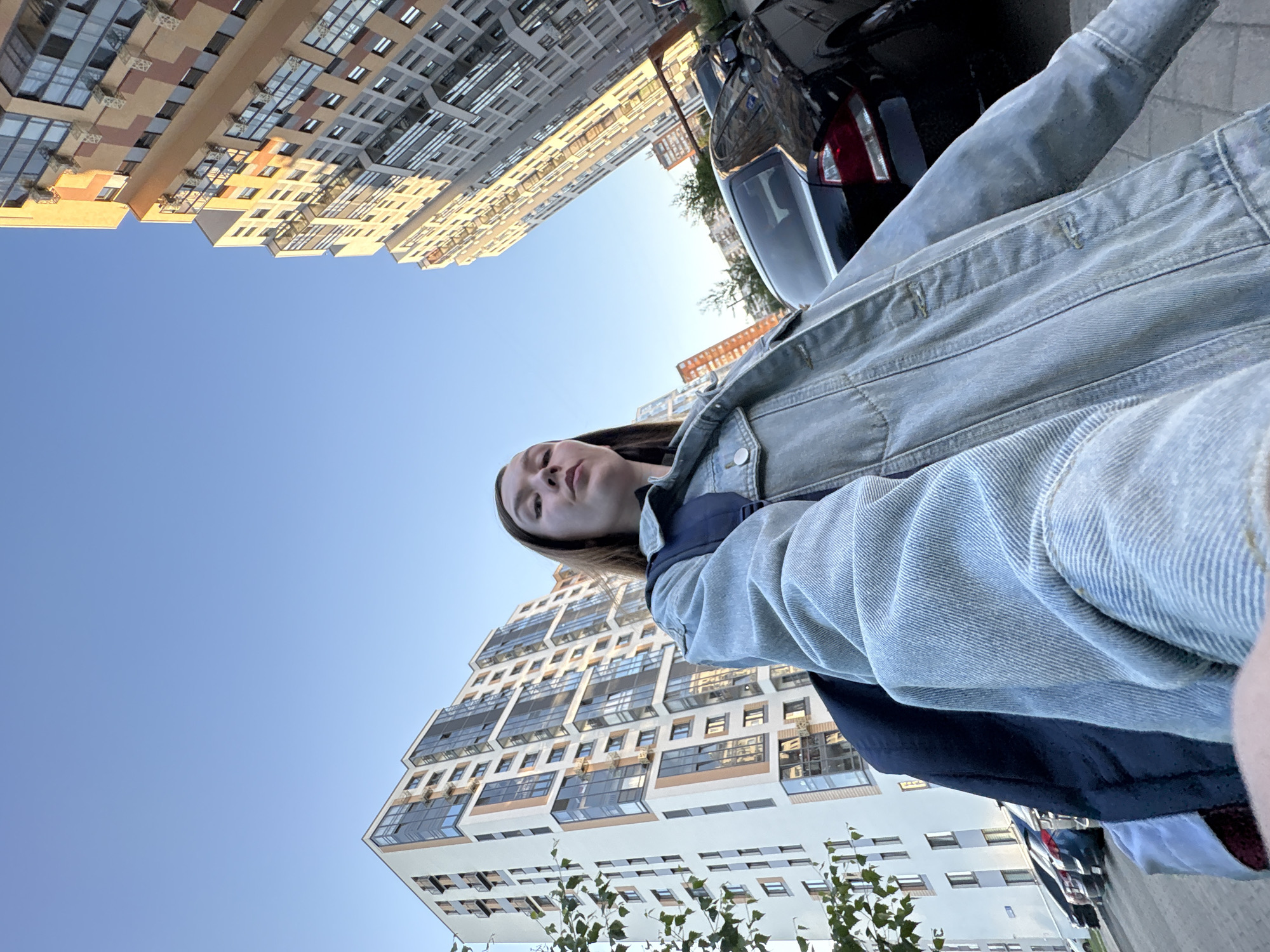 In Izhevsk, Russia, a young woman is taking a selfie outdoors, standing in a paved area with parked cars and modern apartment buildings in the background. She is wearing a light blue denim jacket over a dark top and a backpack. Her face is turned slightly towards the camera, and her expression is neutral. The buildings, characteristic of contemporary urban development, display a mix of light-colored facades and glass windows, with some showing signs of window air conditioning units. The sky above is clear and blue, indicating fair weather. The angle of the shot, looking up towards the woman and the buildings, creates a sense of scale and perspective.