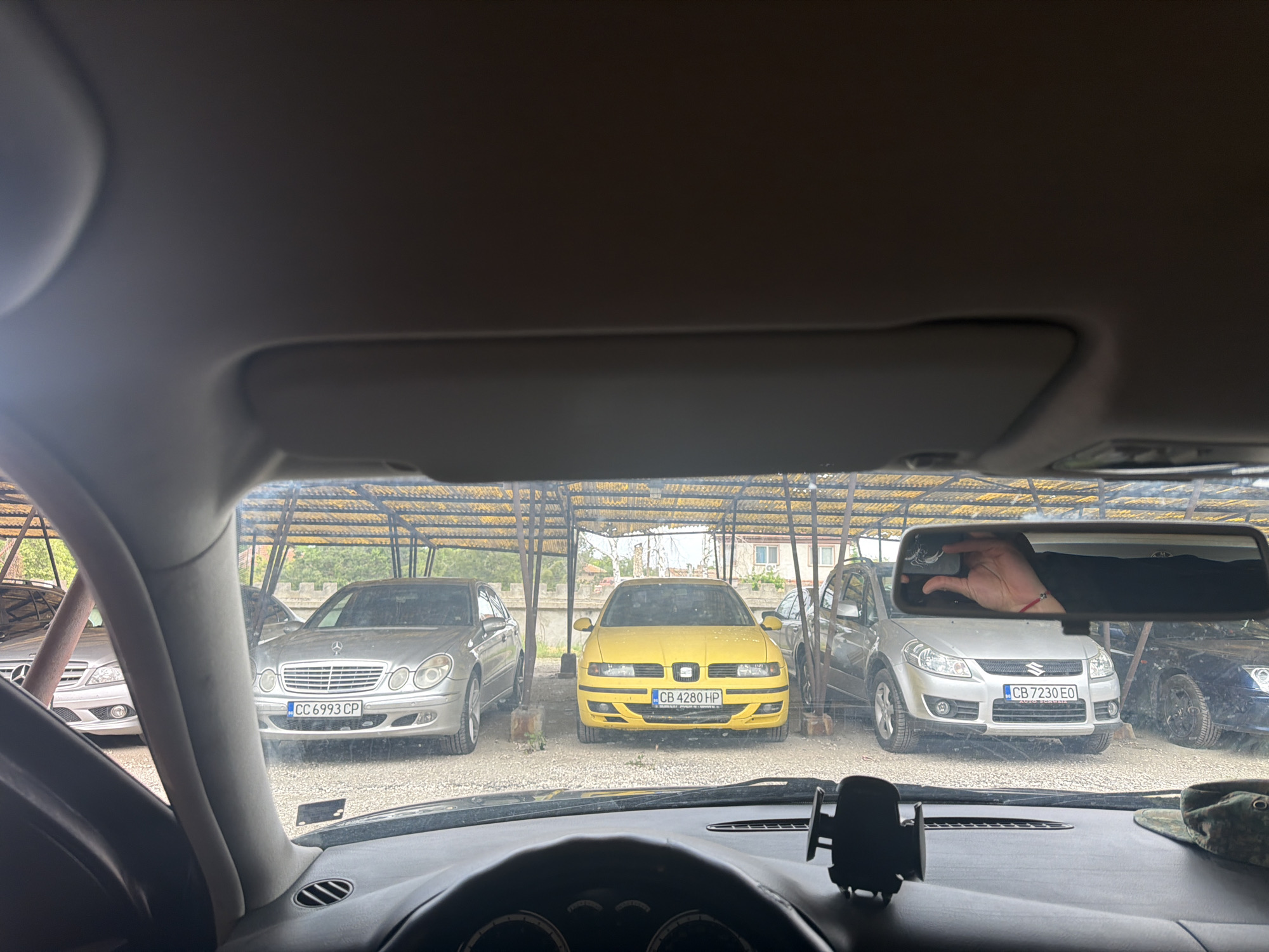 Captured from within a car, the image presents a view of three vehicles parked under a metal canopy, plausibly in Dolna Mitropolia, Bulgaria, based on the Bulgarian license plates: "CC 6993 CP" on a silver Mercedes-Benz, "CB 4280 HP" on a yellow SEAT, and "CB 7230 EO" on a silver Suzuki SX4. The photo is taken from the driver's seat, evidenced by the steering wheel and dashboard. A smartphone mount is on the dashboard. A hand holding a phone is reflected in the rearview mirror. The cars are parked on a gravel surface with vegetation visible in the background. The environment suggests a parking area or lot, possibly at a car dealership or storage facility.