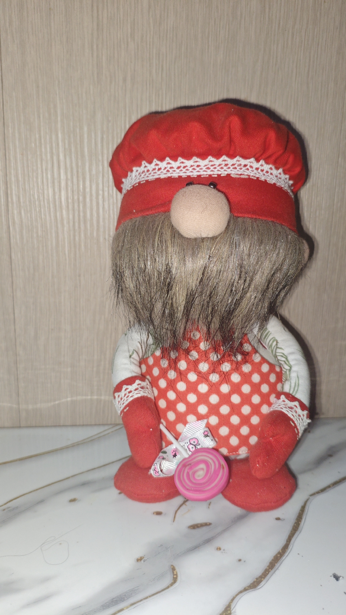 A plush gnome toy is positioned against a wooden plank wall, with its lower half resting on a marbled surface. The gnome wears a red chef's hat with white lace trim and a red and white polka dot apron. Its arms are red, and it is holding a pink and white swirled lollipop. The gnome has a large, shaggy brown and gray beard and a rounded beige nose. The background is a light brown wooden wall with vertical planks. The foreground features a white and gray marbled surface with gold veining. The lighting is even, suggesting an indoor environment. The scene is static, with no visible action or interaction. The location is not specified within Serebriansk, Kazakhstan; this is a generic indoor setting.