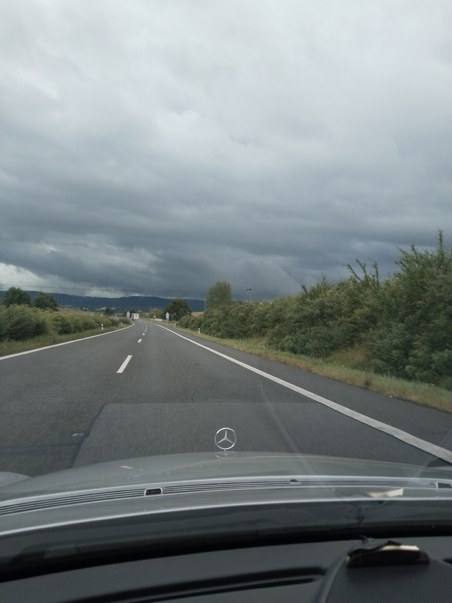 The image shows a view from inside a vehicle driving on a highway. The sky is heavily overcast with dark, stormy clouds. The road ahead curves to the left, with white lines marking the lanes. On either side of the road are dense green bushes and trees. In the distance, there are rolling hills or mountains. The hood of the car is visible in the foreground, with a Mercedes-Benz logo on it. The scene appears to be a highway in or near the city of Husa, Czechia, given the general landscape and infrastructure. There are no people or other vehicles clearly visible in the immediate foreground or on the road ahead. The overall impression is of a journey during inclement weather.