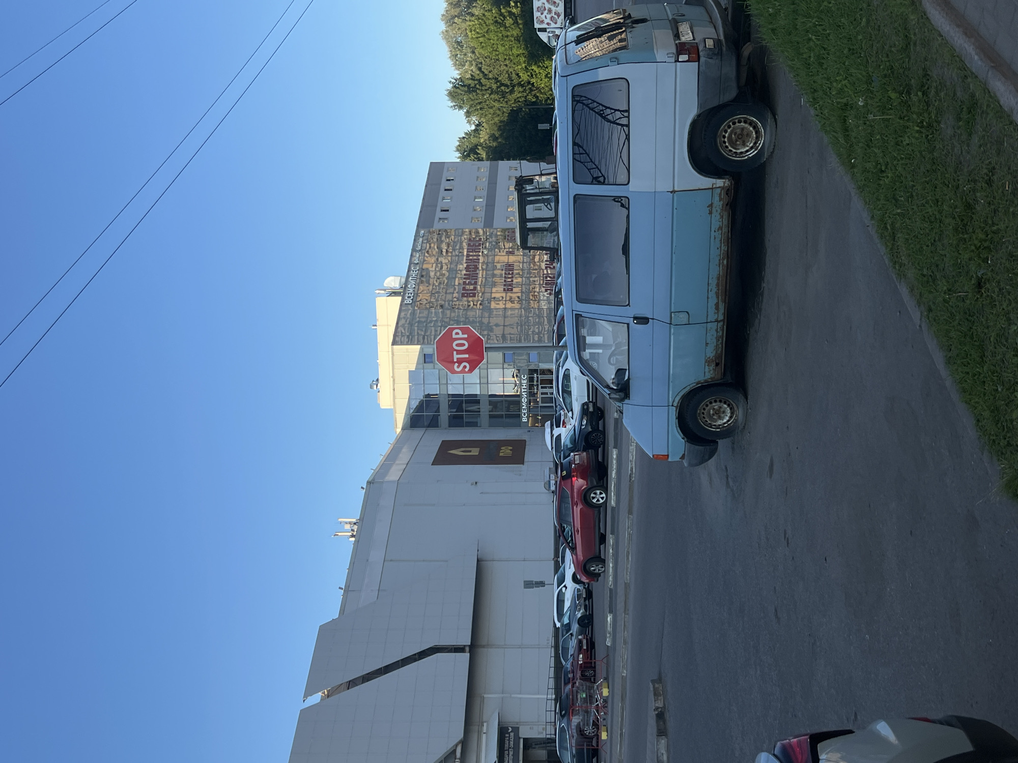 This image captures an outdoor scene at the "Bogatyr" shopping complex, located at Levashovsky Prospekt 18 in Saint Petersburg, Russia. The time appears to be late afternoon or early morning, under a clear blue sky.
The prominent feature is a large, multi-story commercial building with a light-colored facade. On the left section of the building, partially visible text reads "ТВОЯ ИДЕЯ ТОВАРА В ИНТЕРНЕТ-ЗАКАЗЕ" (Your product idea in online order). The central section displays a brown house-shaped logo with the text "ПРО" below it, identifying it as a "Petrovich" home improvement store. The right section features a reflective glass facade with "ВСЕМИРНЫЙ ФИТНЕС" (Vsemirny Fitness), "БАССЕЙН" (Pool), and a partial phone number (+7 (812) 24...) visible. A red octagonal STOP sign stands in front of this reflective facade. Overhead utility lines are visible crossing the upper right portion of the sky.
In the foreground, a heavily rusted and two-tone (light blue front, lighter blue/grey rear) Volkswagen Transporter T4 van is parked facing right, with its rear driver's side tire deflated. The bottom half of the van, particularly the driver's side, shows extensive corrosion. To the left, the rear bumper and taillight of a dark-colored car are partially visible.
The midground features a large asphalt parking lot with numerous parked vehicles, including a red SUV (Toyota RAV4 style), various sedans, and SUVs in white, black, and grey. Shopping carts are clustered near the left edge of the parking area. Behind the van, a yellow and black construction vehicle (possibly a backhoe or front-end loader) is partially obscured. Green grass lines the curb along the right side of the foreground, and a dense line of green trees is visible behind the buildings on the right side of the image. No people are discernible in the scene.