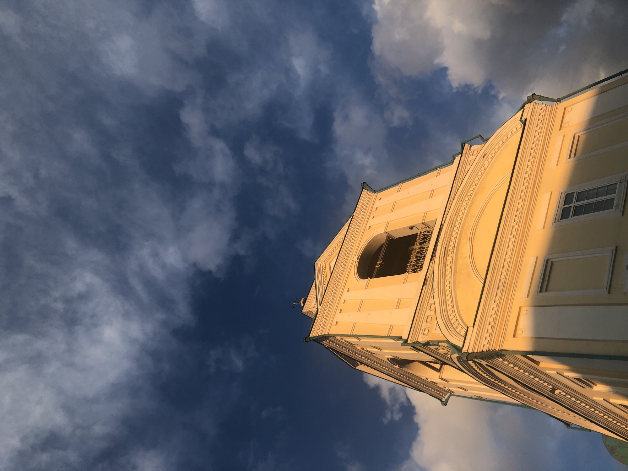 The image shows a low-angle view of a yellow building under a sky with mixed dark blue and white clouds. The building appears to be a church bell tower, featuring architectural details such as arched windows, decorative moldings, and a cross at the top. The setting sun casts a warm light on the building's facade, highlighting its texture. The location is in Krasnoyarsk, Russia.