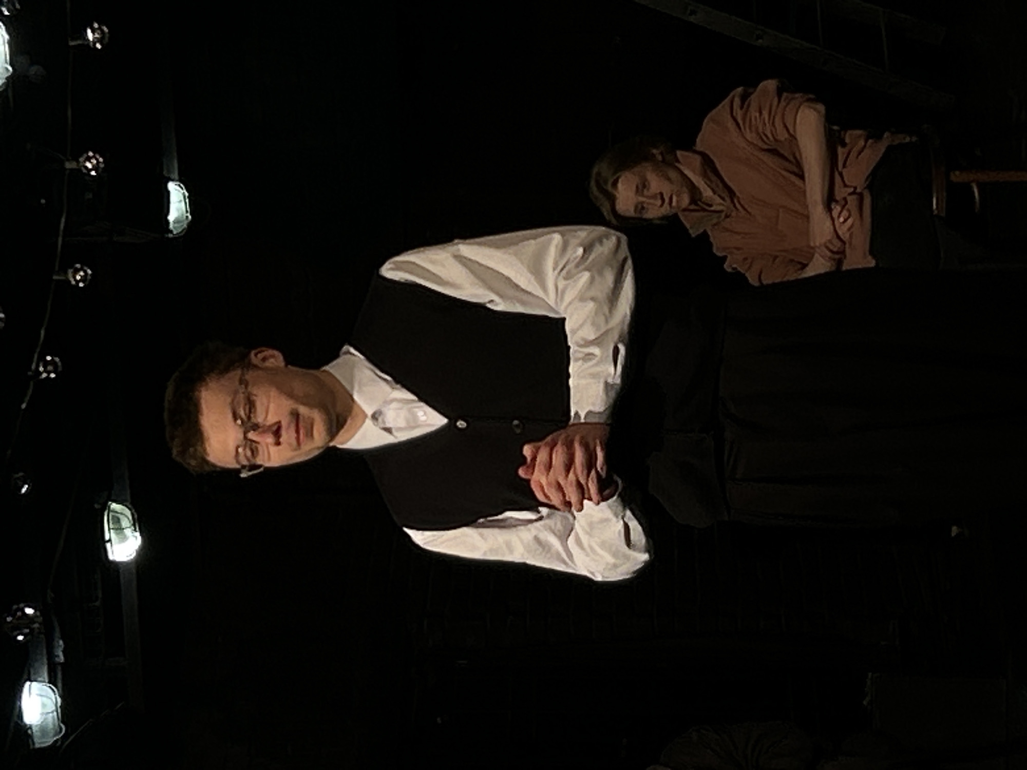 The image depicts two men within a dark setting, characteristic of a theater stage or industrial space, with the photograph itself rotated 90 degrees counter-clockwise. In the foreground, a man with short brown hair and glasses is positioned in the lower half of the frame. He wears a white collared shirt and a black vest, with his hands clasped together at his waist, looking forward with a neutral, contemplative expression.
On the left side of the scene, several industrial-style light fixtures, each with a protective cage, are arranged vertically and emit a soft, localized glow. In the upper right portion of the background, a second man is visible, appearing to lean forward over a dark, indeterminate surface. He wears a light brown, long-sleeved shirt, and his expression appears serious.
The overall environment is predominantly dark, with the subjects highlighted by focused illumination. No specific activity beyond posing or performance is clearly indicated. No text is visible in the image. The specific location and time of day are unknown, given the artificial lighting and dark background.