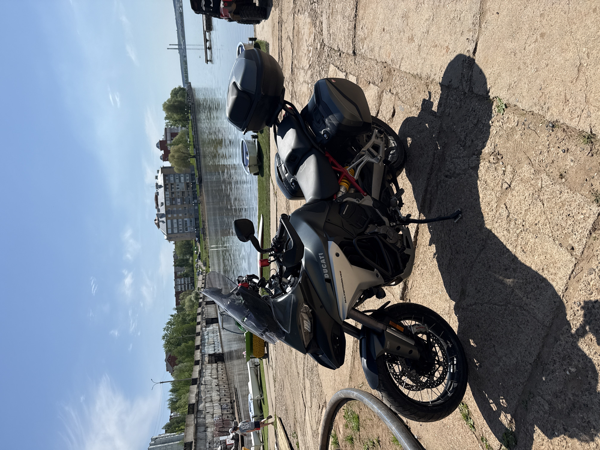The image shows a Ducati Multistrada V4 S motorcycle parked on a concrete surface next to a body of water. In the background, there are buildings and a bridge. Based on the urban environment and the presence of the Votkinsk Hydroelectric Dam and P. I. Tchaikovsky Museum complex (likely indicated by the bridge and buildings along the water), the location is likely in or near Votkinsk, Russia. There are hard panniers and a top box attached to the motorcycle, indicating it is set up for touring. The motorcycle is parked on its kickstand, and its shadow is visible on the concrete. There are no people clearly visible.