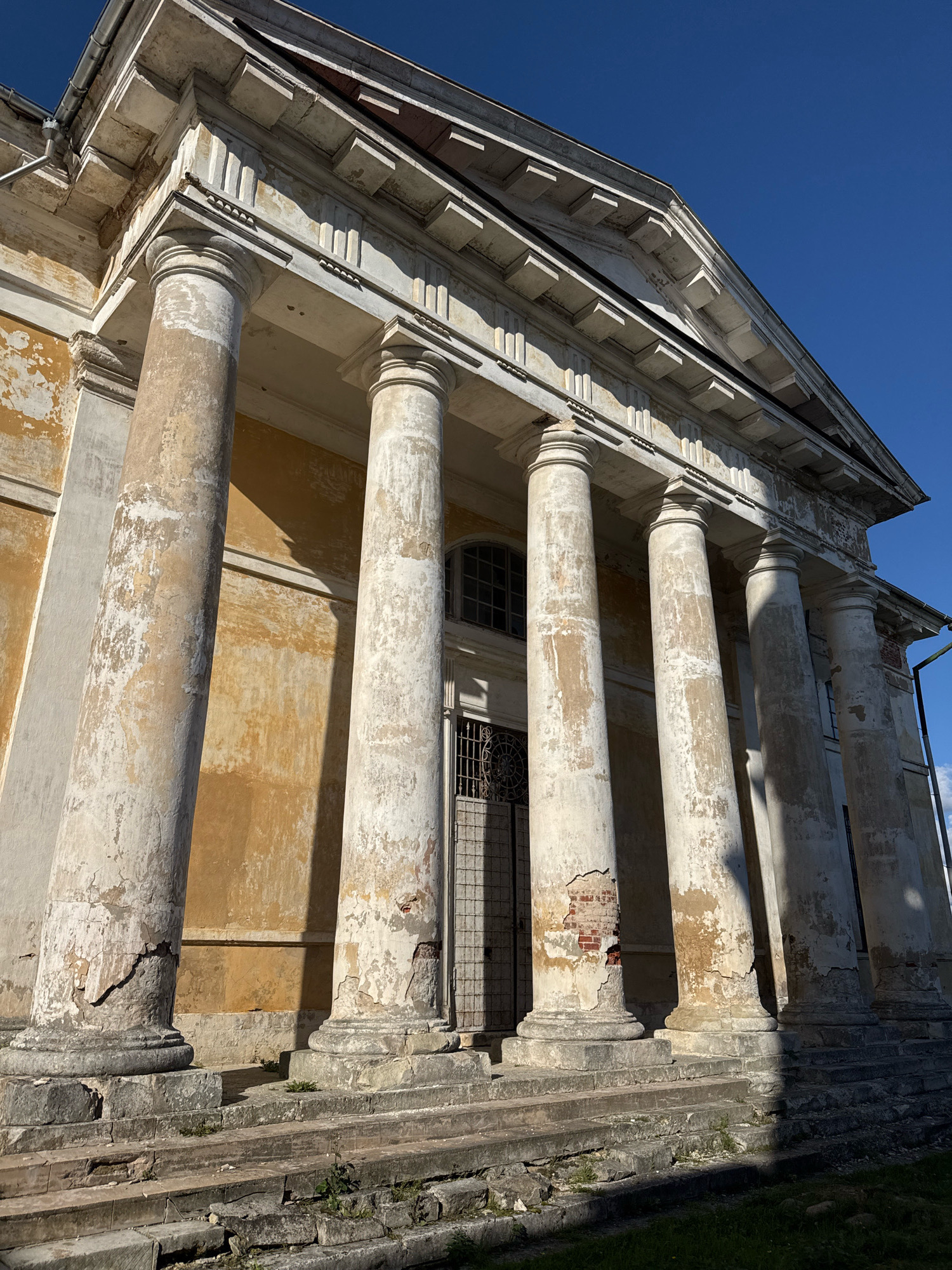 The photograph captures a sunlit view of the facade of the Borisoglebsky Monastery in Torzhok, Russia. The image focuses on a portico with six large, weathered columns that exhibit significant signs of decay, including peeling paint and visible brickwork. The columns are set against a pale yellow, aged stucco wall, which also shows discoloration and wear. Above the columns, an architectural entablature with decorative elements and a triangular pediment is visible. Sunlight casts strong shadows across the columns and the building's facade, creating a stark contrast with the deep blue sky. In the background, a metal-grated door and a window with a lattice are partially visible. The foreground shows a worn, stone staircase leading up to the portico, with patches of green grass and scattered debris. The overall impression is one of a grand, historic structure in a state of disrepair, bathed in bright daylight.